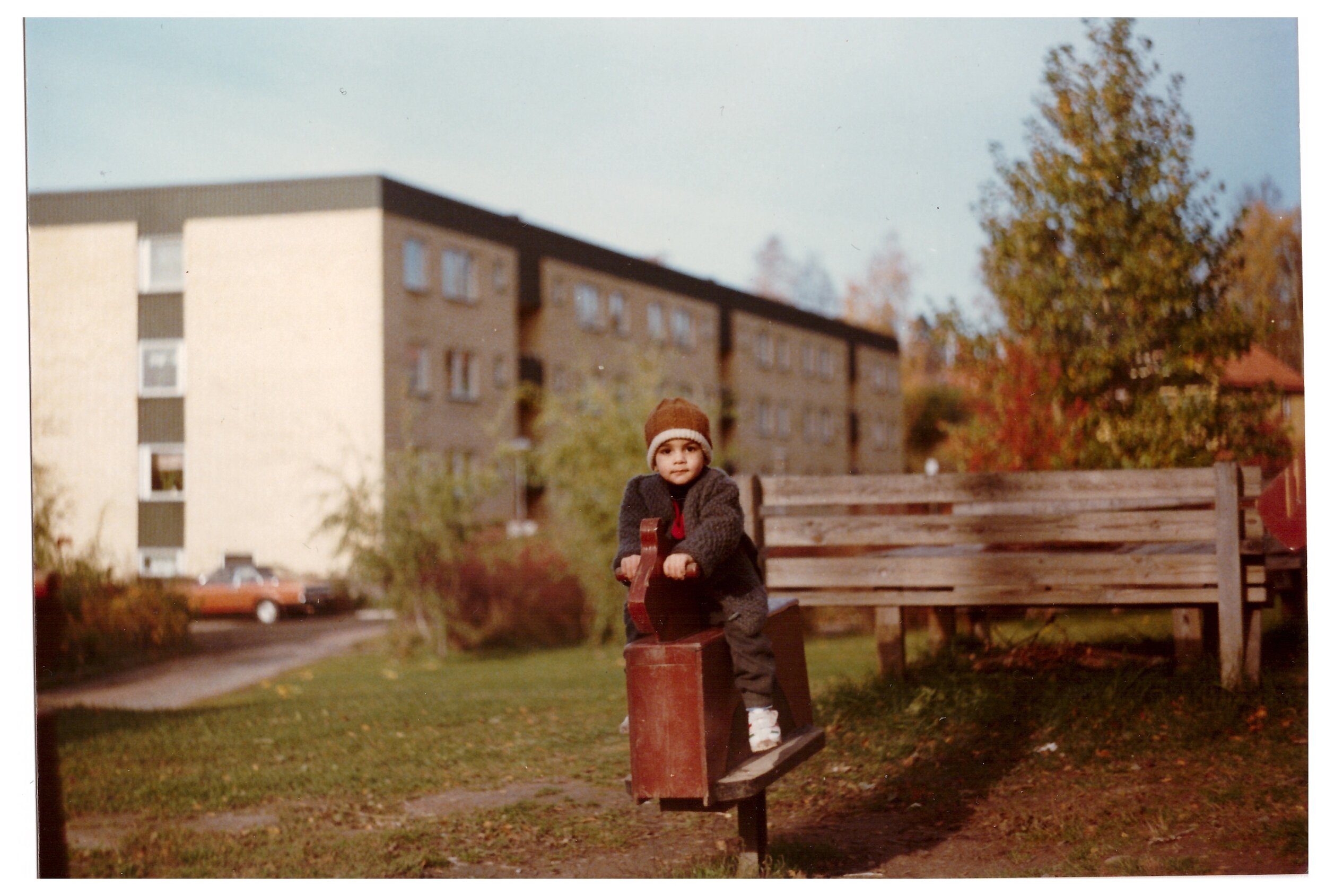 Child playing outside on a wooden seesaw in a park, with apartment buildings and trees in the background.