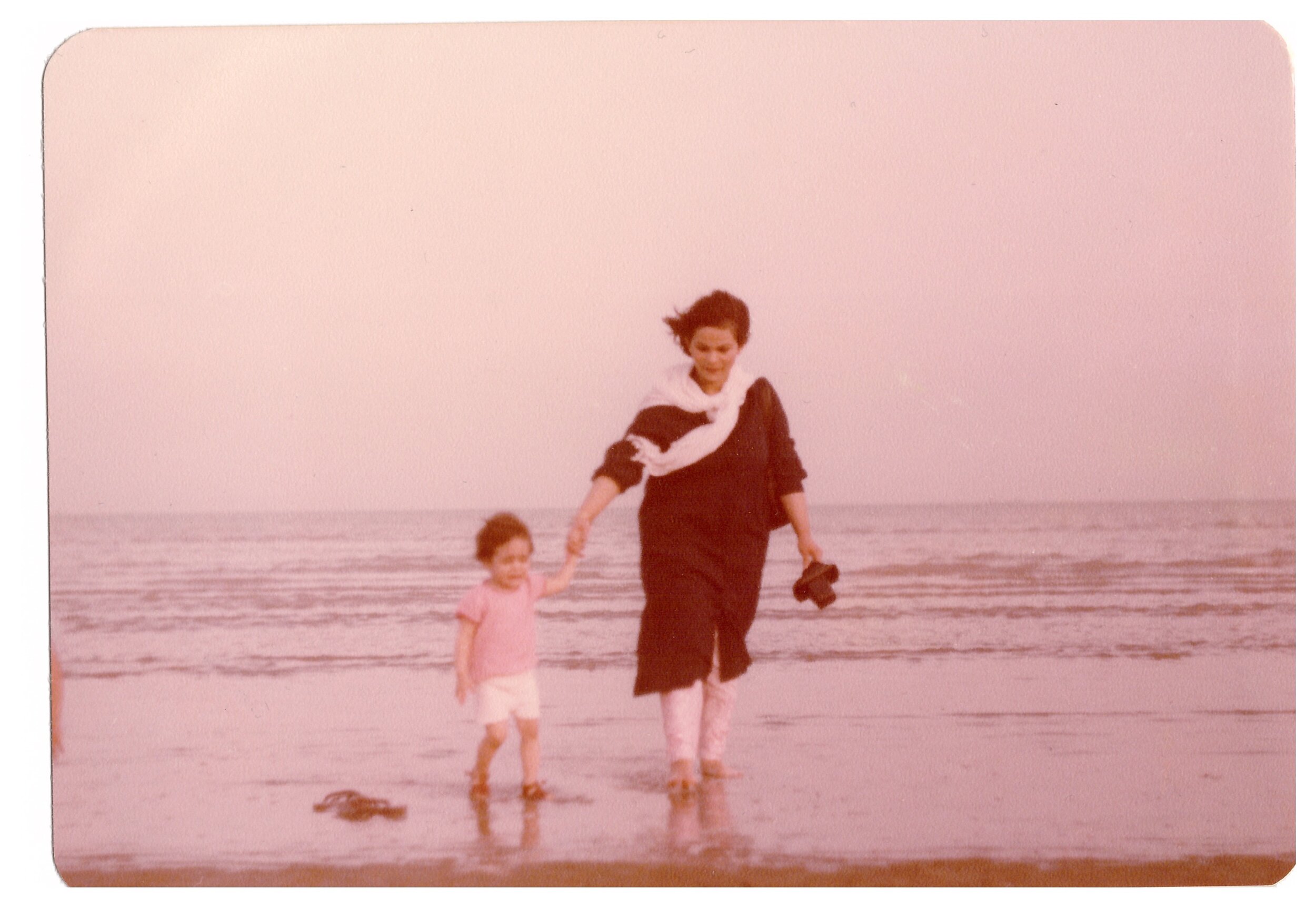 Woman and child holding hands, walking on a beach with water waves in the background.