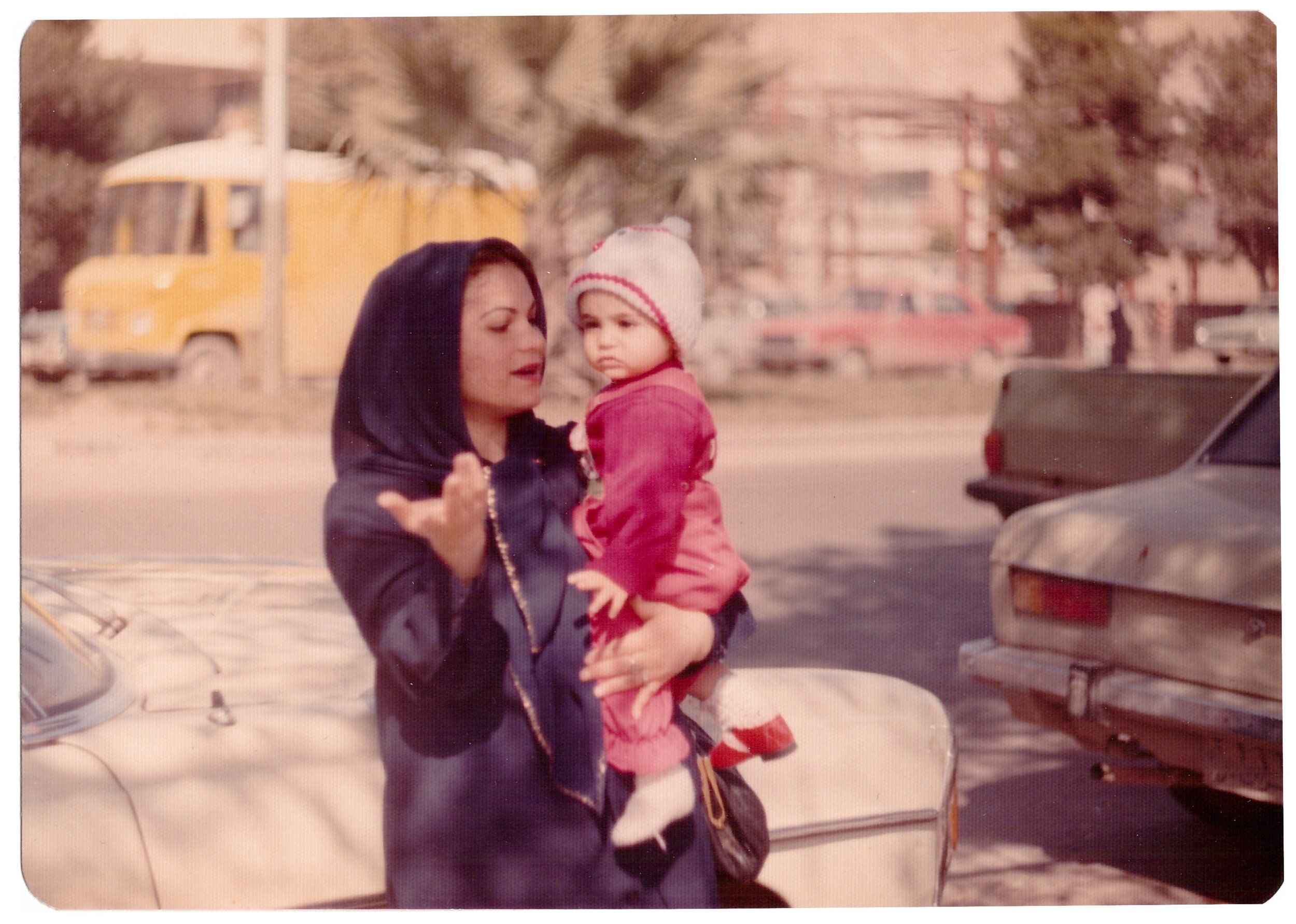 Woman in dark clothing holding a child in pink outfit and hat, standing on a street next to parked cars.