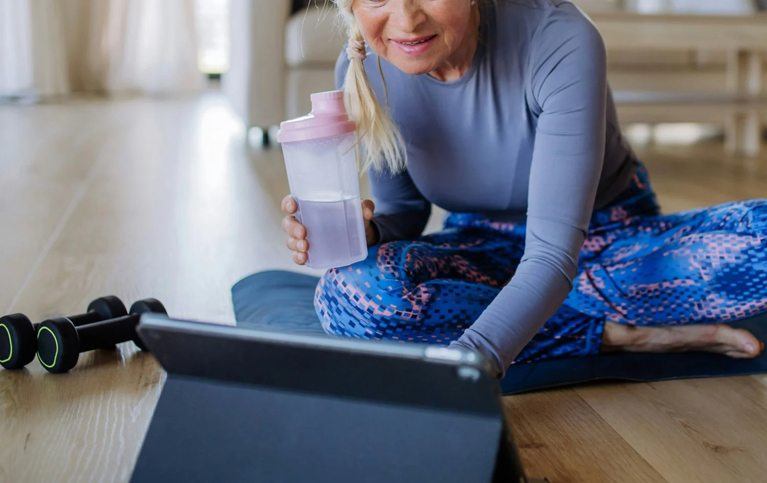 A woman dressed in workout clothes sitting on a yoga mat on the floor, holding a water bottle, with dumbbells nearby, looking at a tablet.