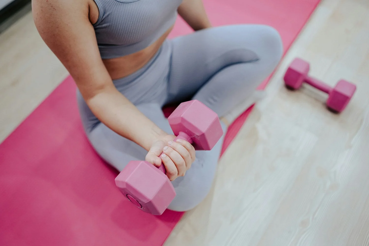 Person sitting on pink exercise mat, lifting pink dumbbells in a fitness setting.