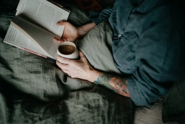 Man laying in bed reading a book with a cup of coffee