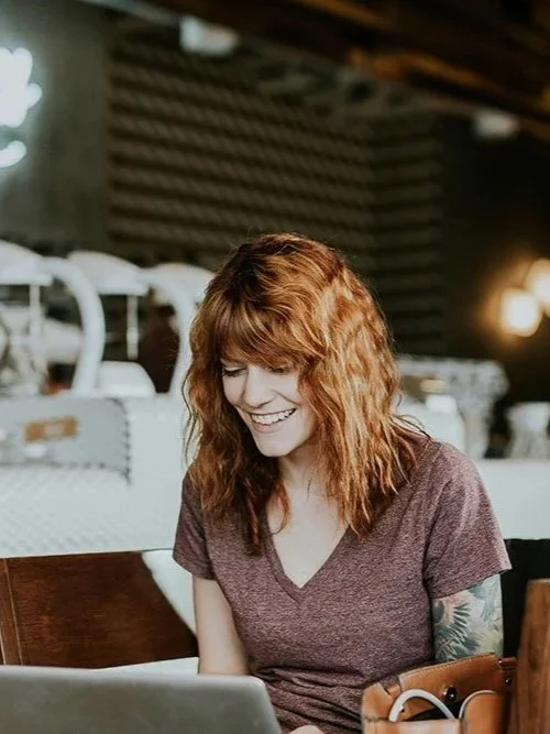 Red haired woman in coffeeshop smiling at laptop