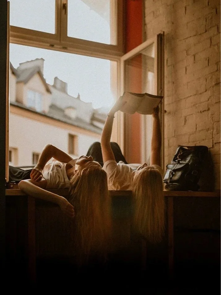 Two female friends reading book in window sill