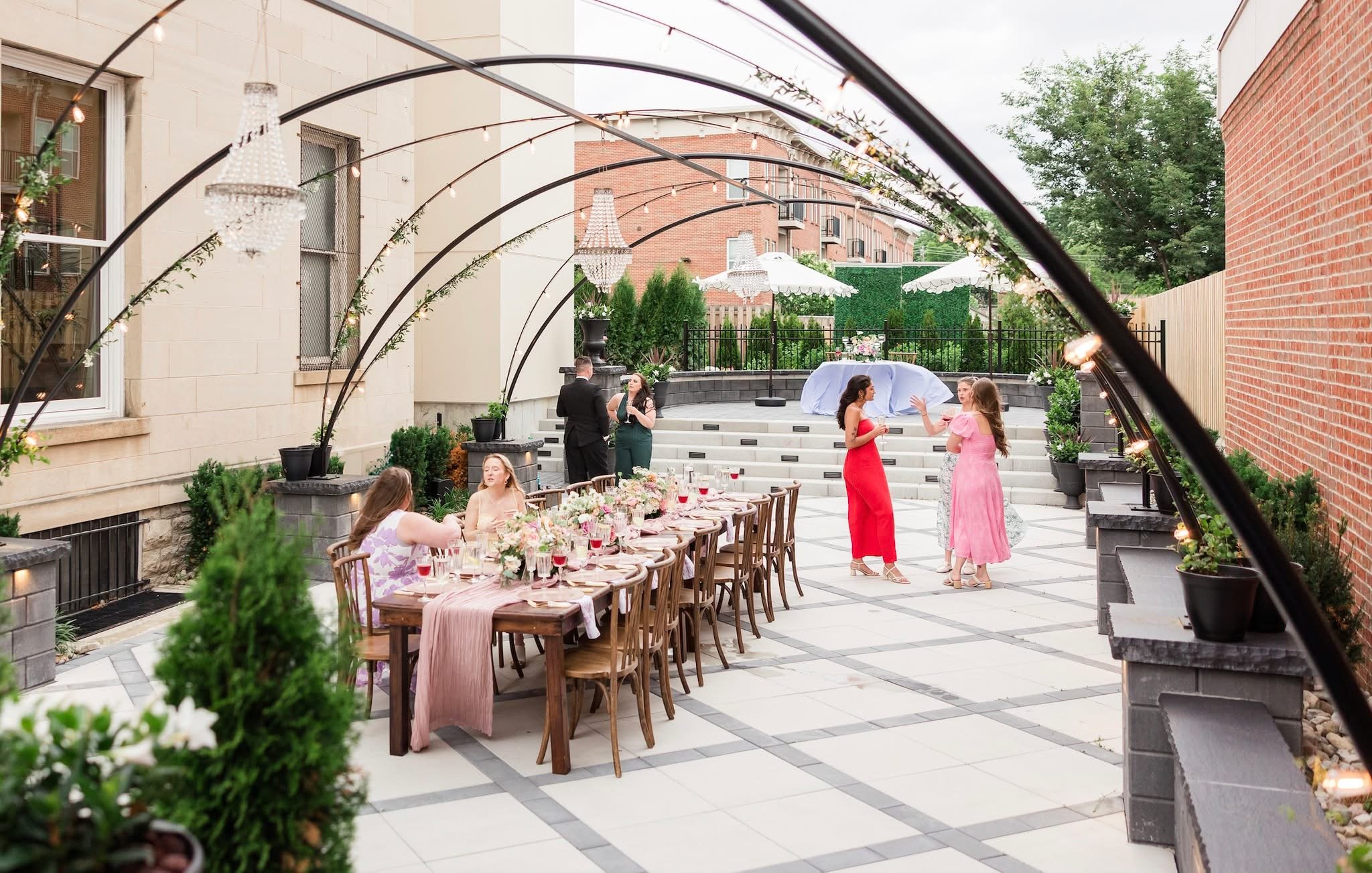 A modern multi-story house with a backyard featuring a pool, hot tub, and lounge area. The backyard is enclosed with black railing and adorned with potted plants and flowers.
