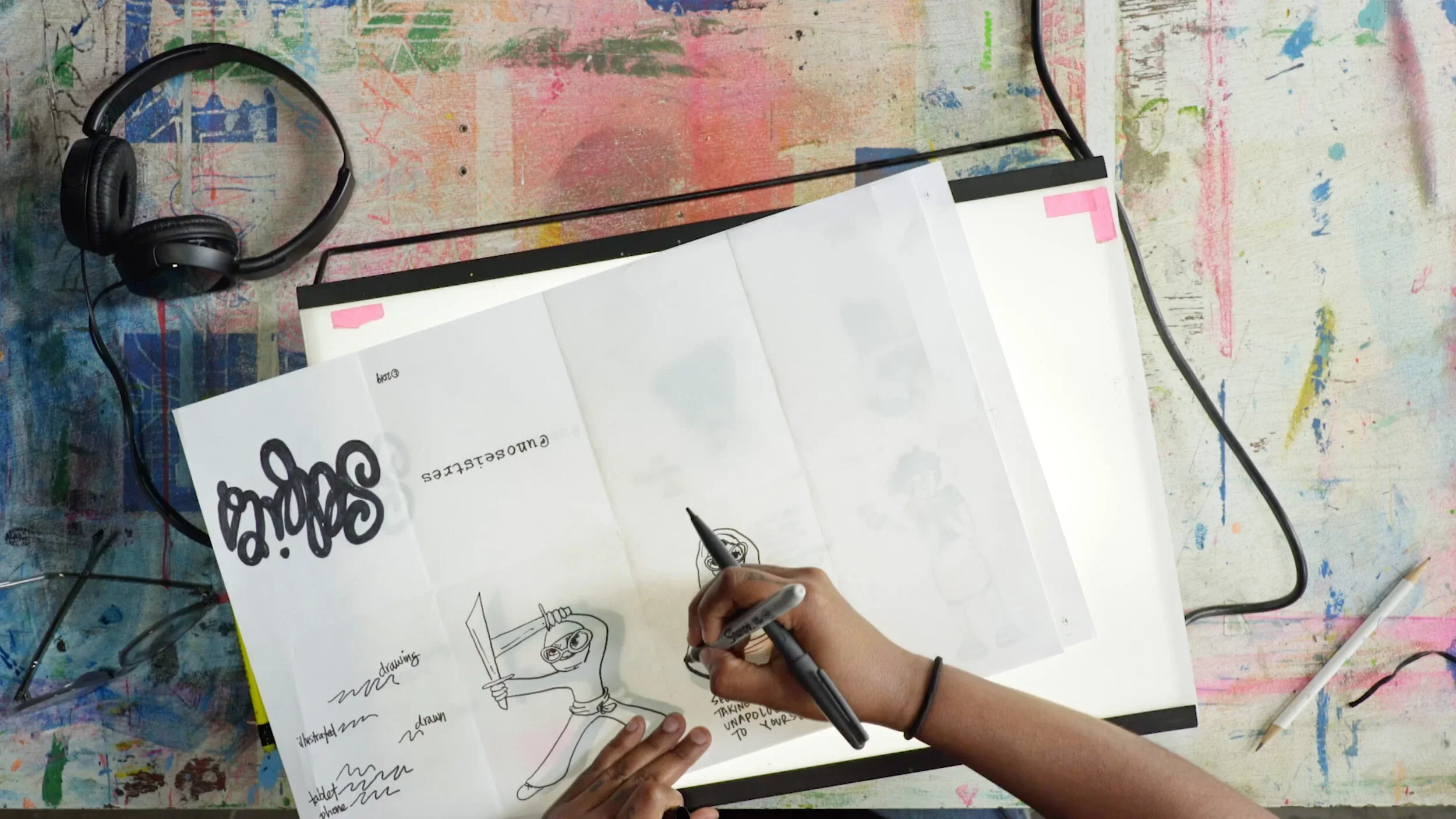An artist sketching a character in a sketchbook at a paint-marked worktable — still from a T Brand Studio video, branded content for Calvin Klein.