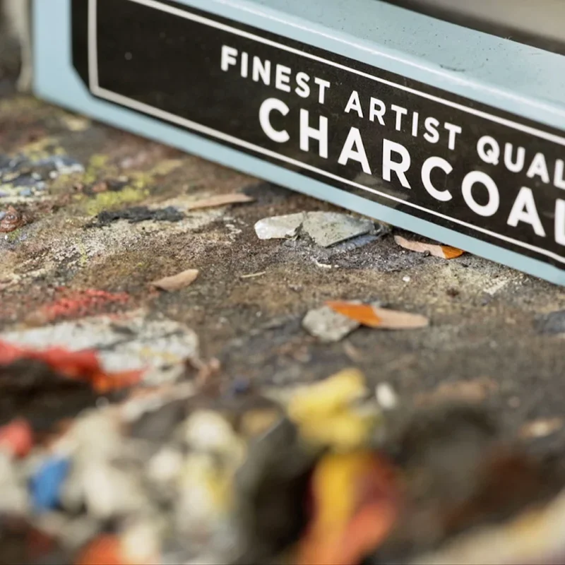 A box of artist's charcoal on a paint-spattered studio floor — Vilcek Foundation artist profile, THEY documentary work.