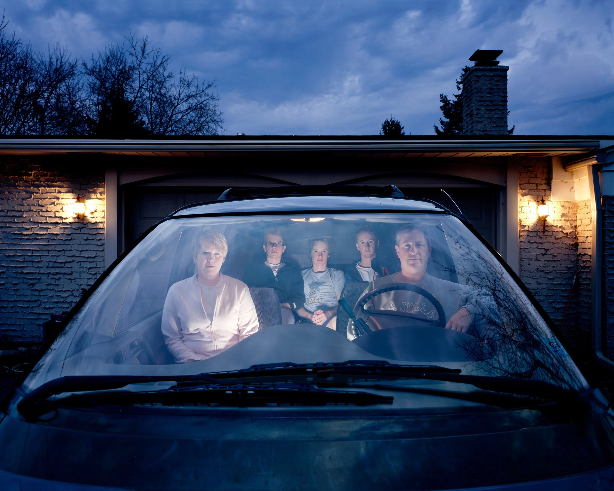 Self-Portrait with Family, 2007 — Julie Mack and her family seated inside a parked car at dusk, photographed through the windshield in front of a suburban brick house.