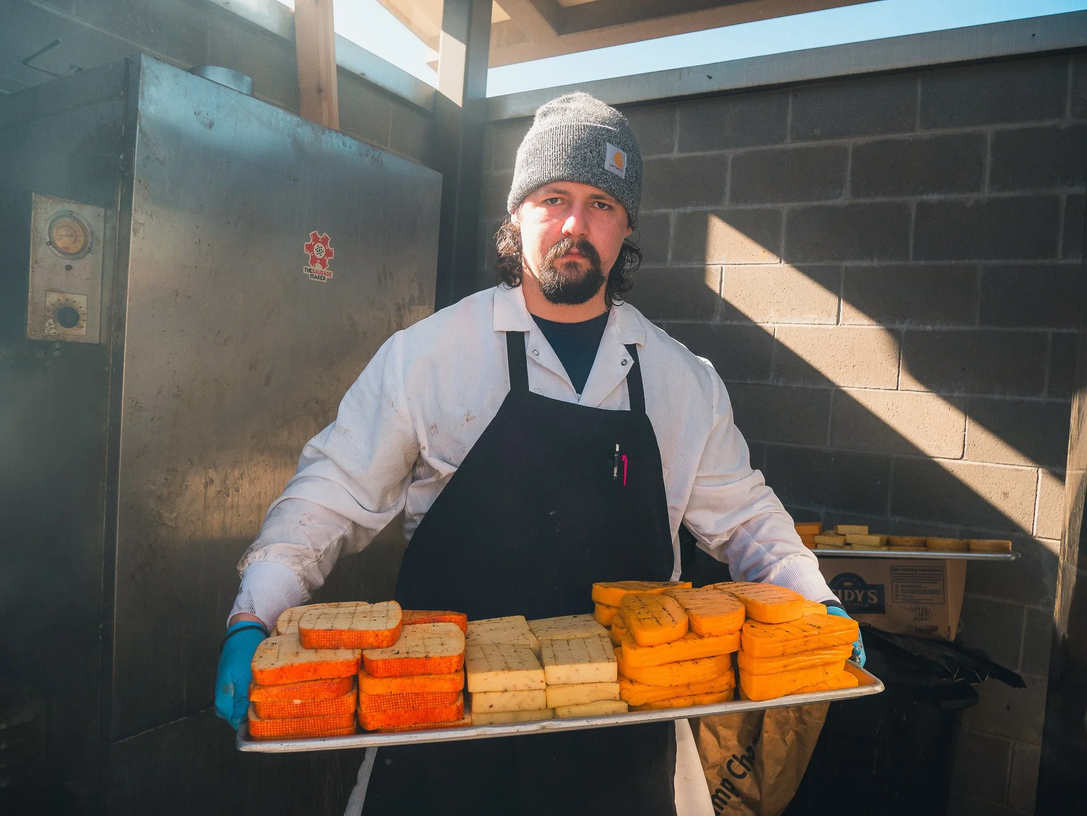 A maker in a beanie and apron behind a tray of smoked cheeses — Teamshares small-business profile.