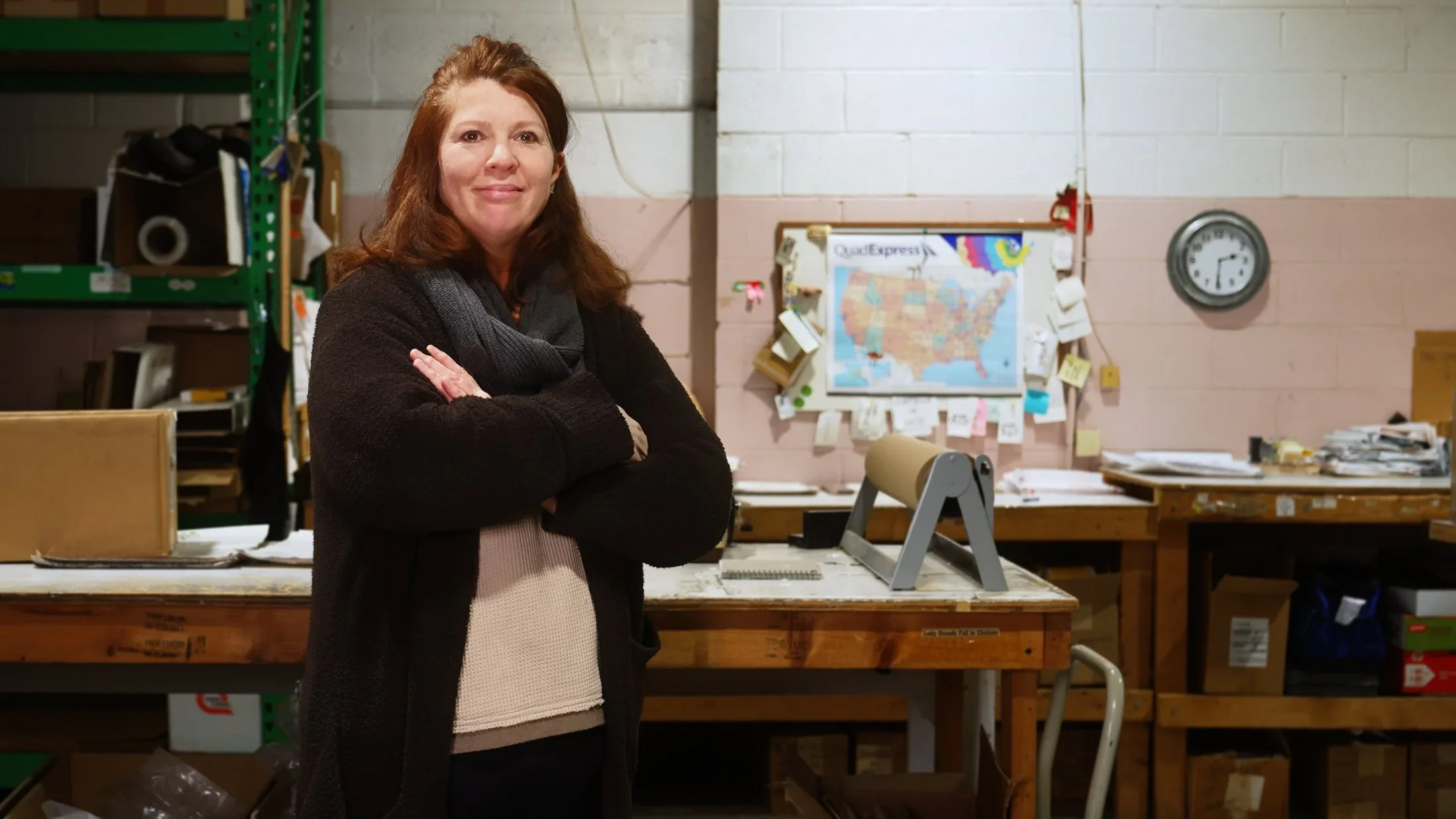 A woman standing arms-crossed in an industrial workspace with a wall map and clock — Teamshares employee-owner portrait.