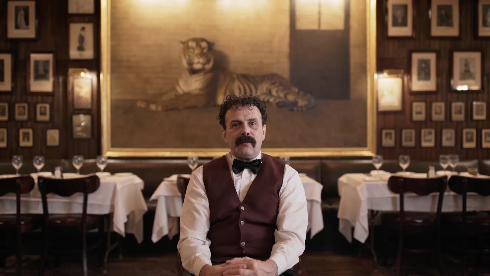 A waiter in a vest and bow tie seated at Keens Steakhouse beneath its framed tiger painting — THEY documentary work.