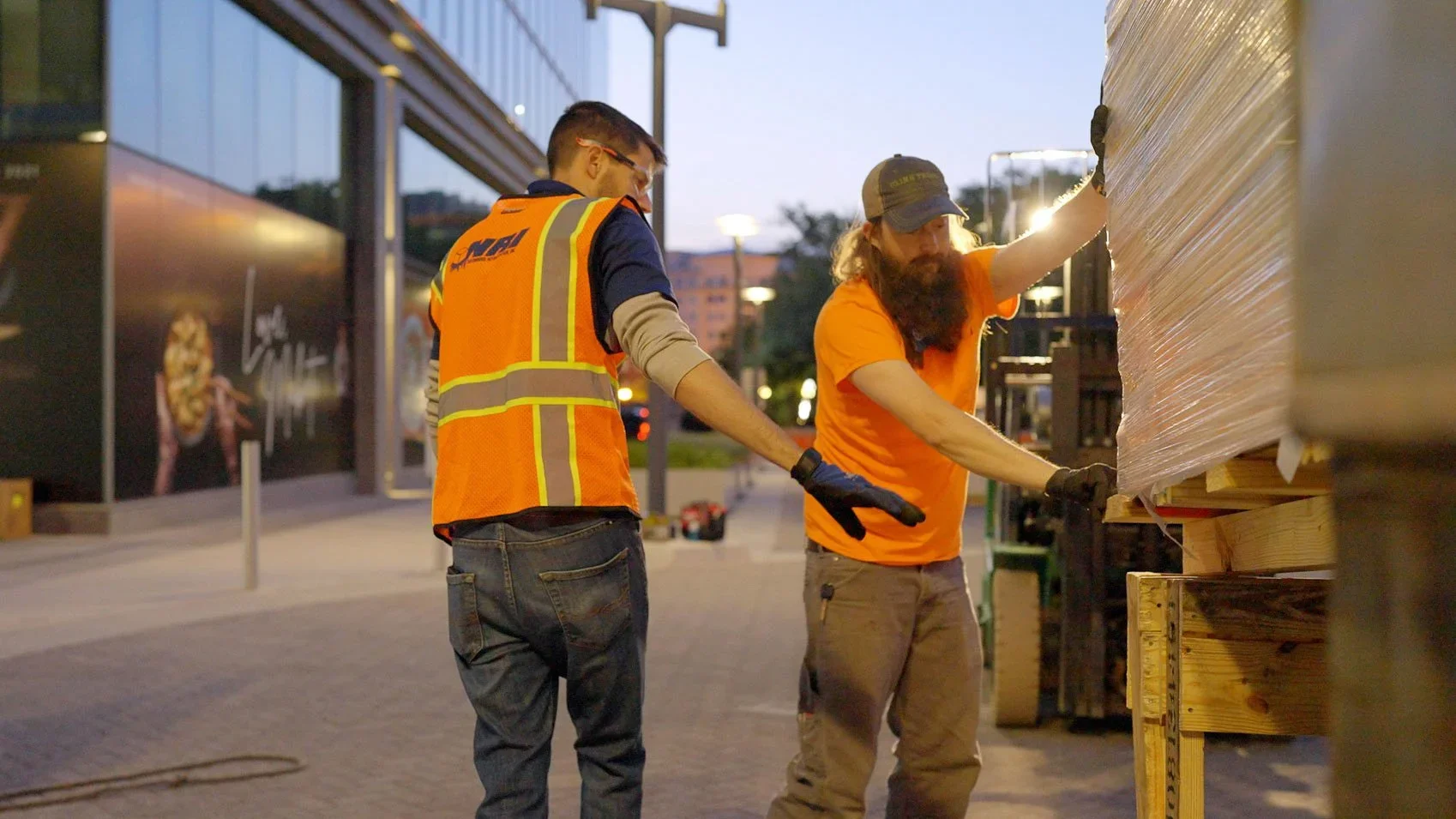 Two workers in orange safety vests handling wrapped pallets outdoors at dawn — Teamshares documentary still for NRI.