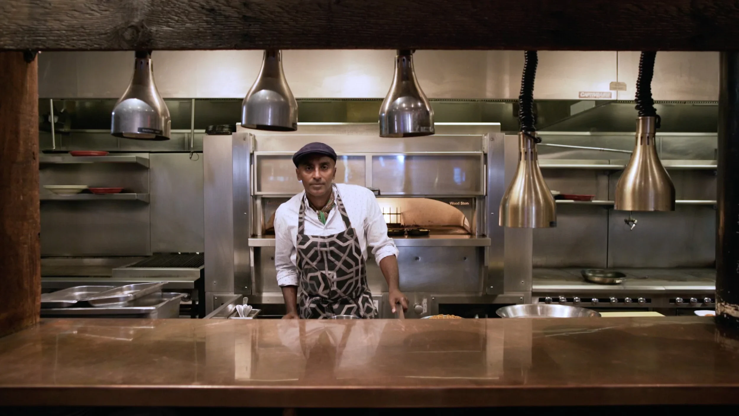 Chef Marcus Samuelsson in a stainless-steel restaurant kitchen under heat lamps — Vilcek Foundation profile, THEY documentary work.
