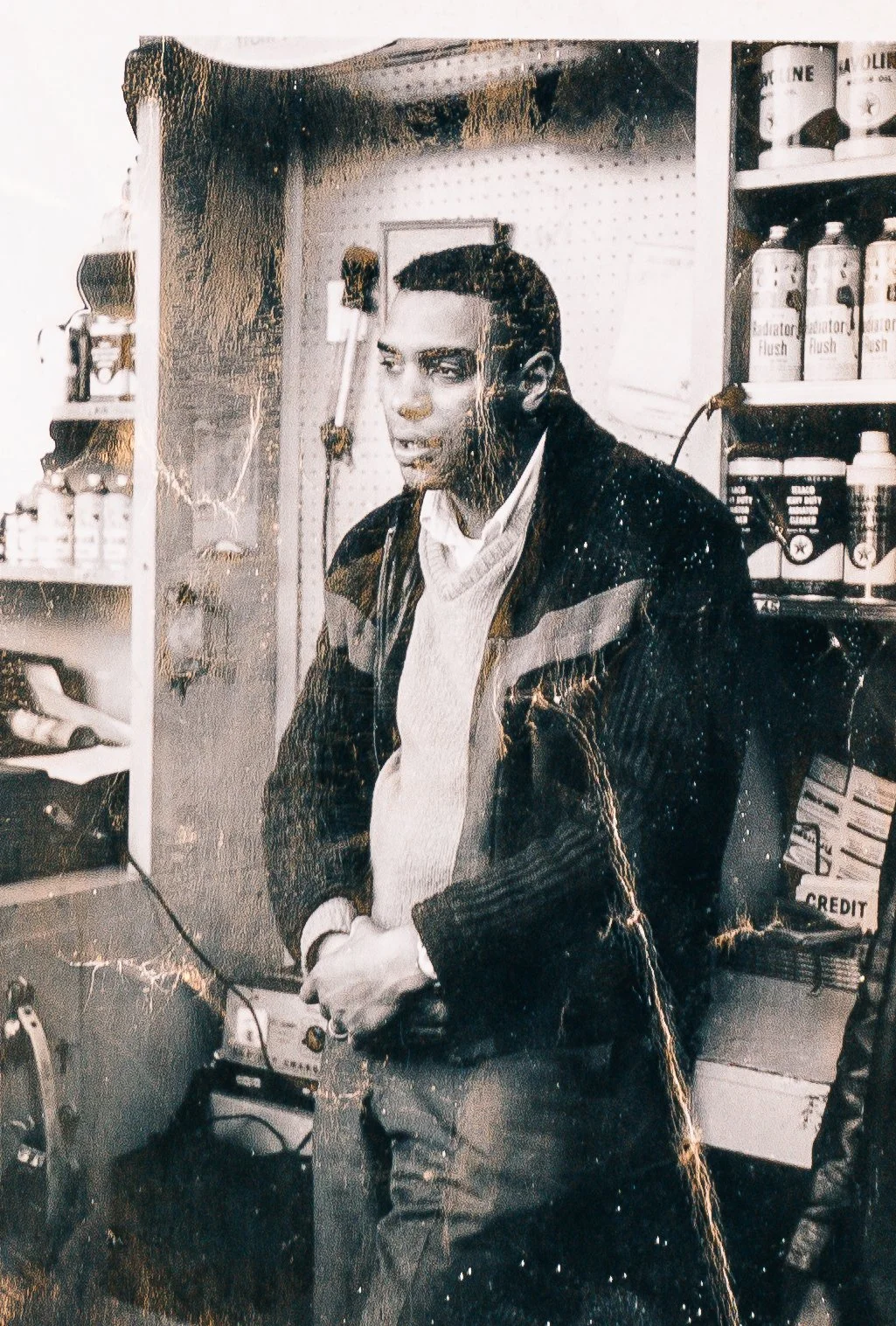 Black and white photo of a man with slicked-back hair wearing a dark jacket over a white shirt, standing inside a store or pharmacy. Shelves with bottles and products are visible in the background.