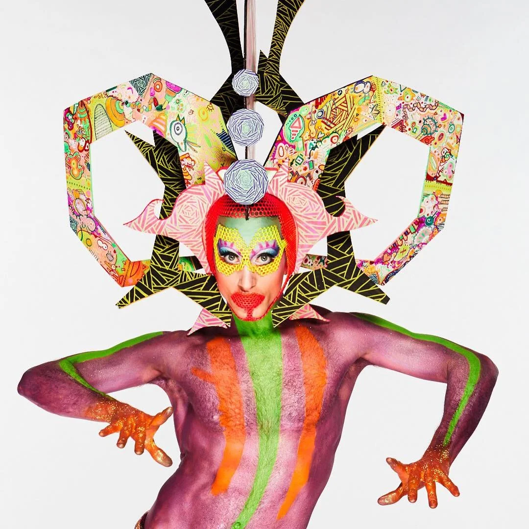 A person with colorful makeup, orange-glitter nails, and body paint is wearing a creative, abstract headpiece with geometric and comic-like patterns. The headpiece features large, three-dimensional shapes and bright colors, resembling a fantastical, 