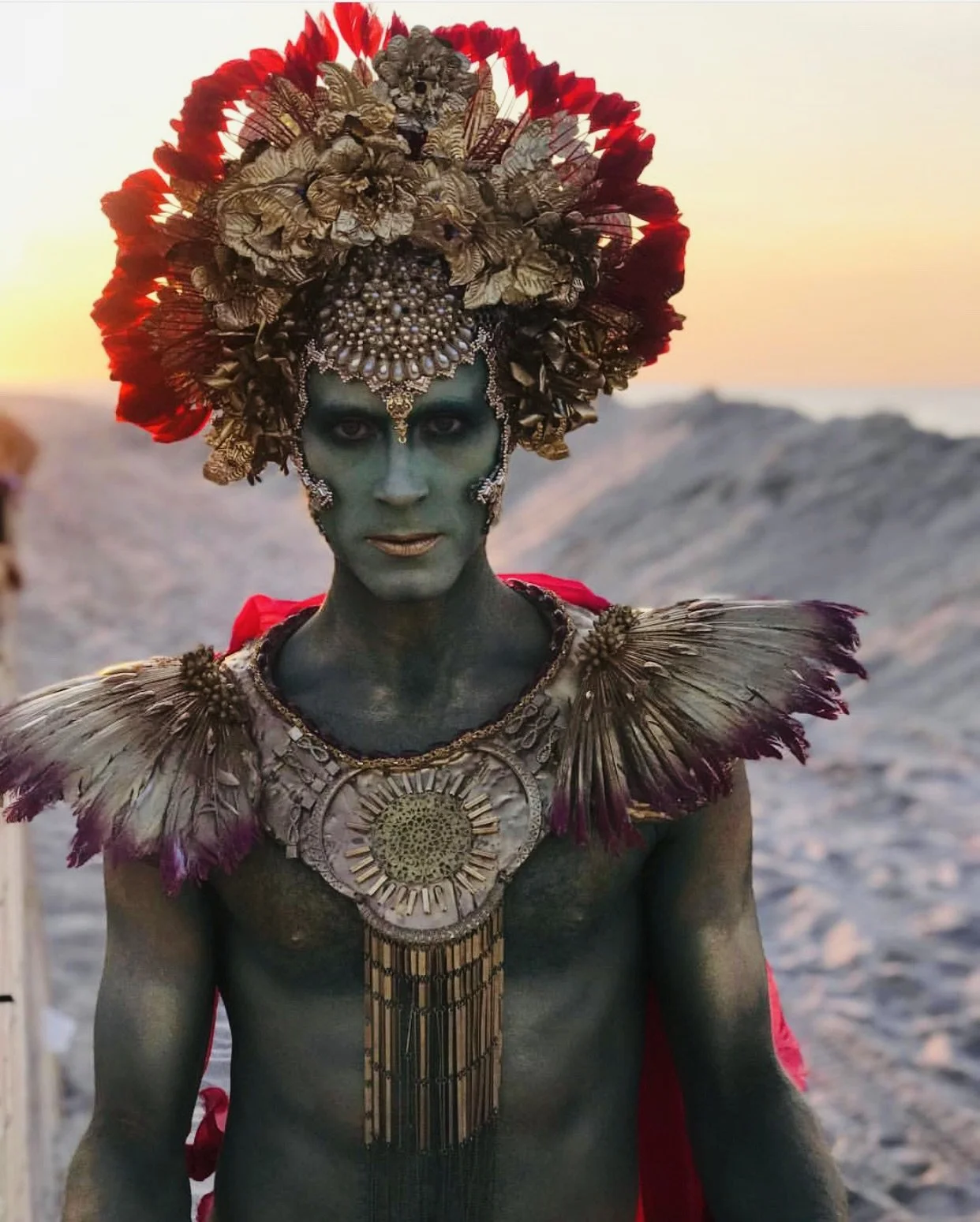 Person with green face paint dressed in elaborate costume with large feathered headdress, ornate jewelry, and feathered shoulder adornments, standing outdoors at sunset.