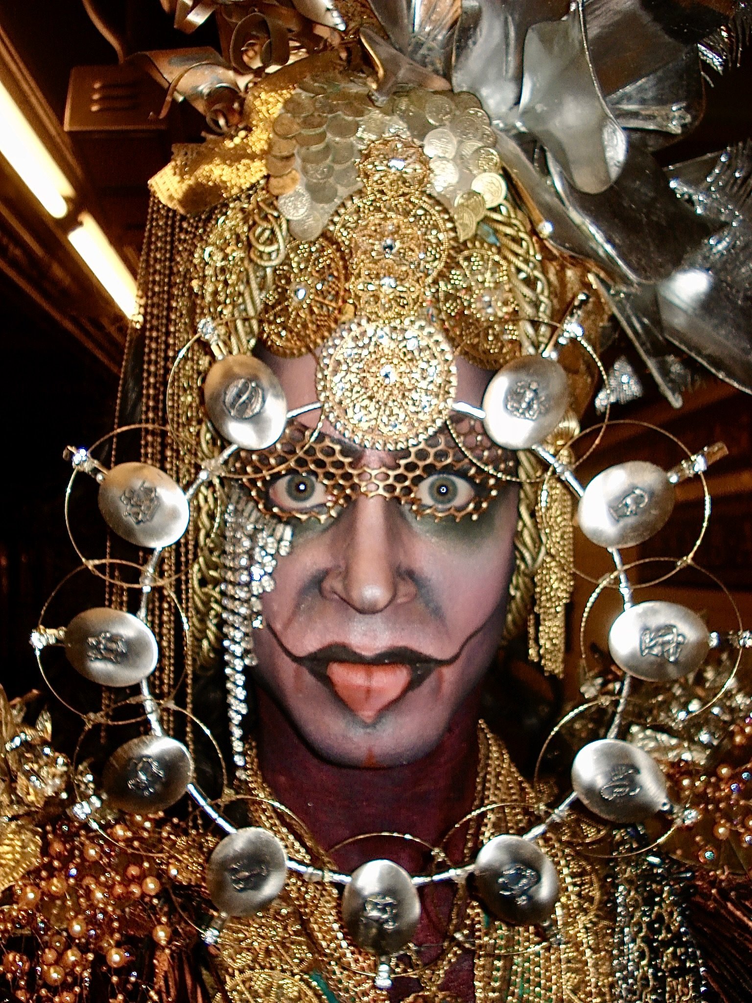 Person wearing elaborate gold costume with a heavily decorated headpiece, face makeup, and jewelry. The headpiece has numerous gold embellishments, coins, and circular ornaments. Face makeup includes dark eye makeup and a painted mouth in a theatrica