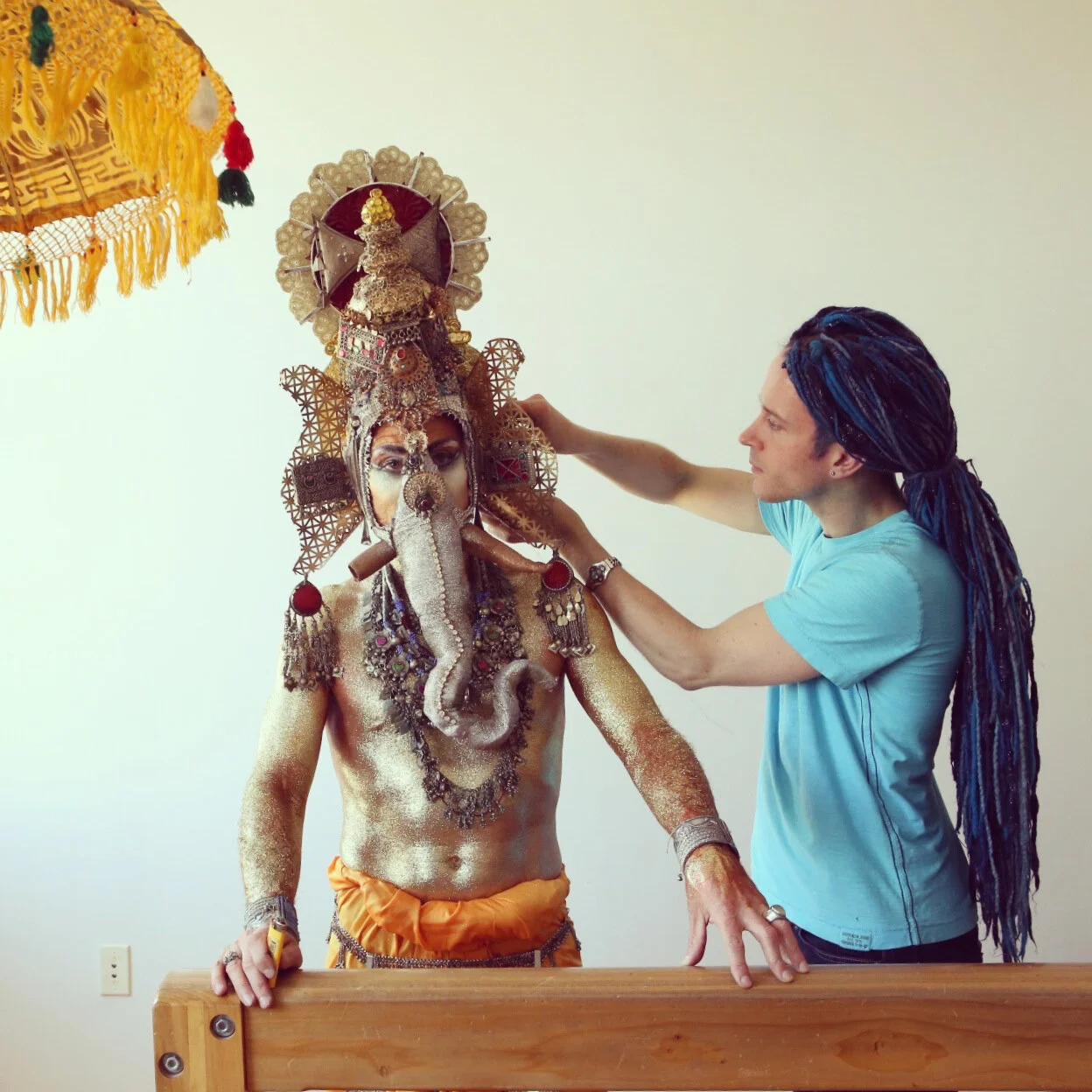 A performer with ornate costume and face paint is being helped by Darrell to adjust his elaborate headpiece. The costume resembles traditional Indian or Southeast Asian deity or mythological figure attire, with jewelry, paint, and headgear.