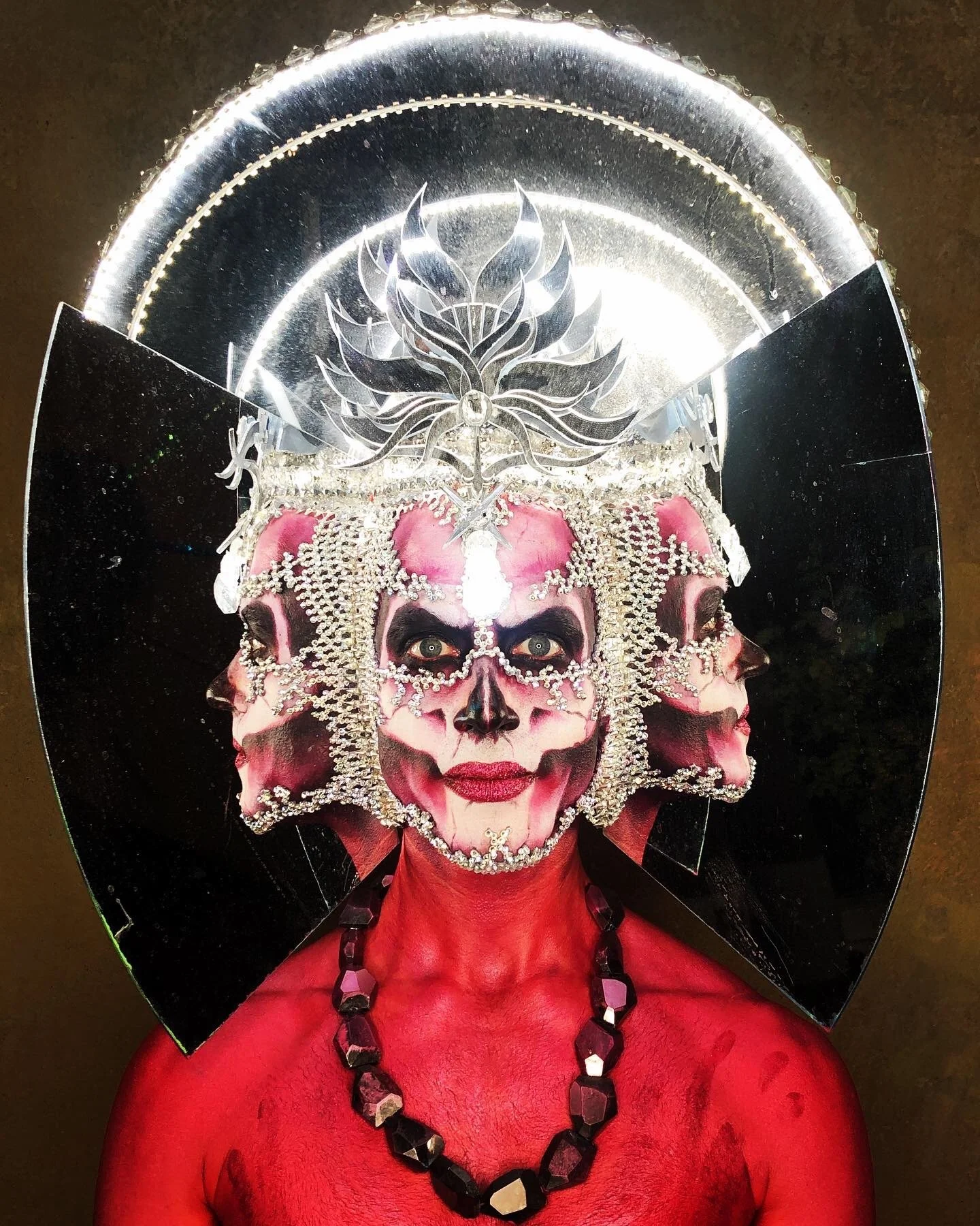 Person dressed in elaborate costume with face painted like a skull, wearing a large ornate headpiece with crystals and a reflective circular element behind, body painted red, and adorned with a black beaded necklace.