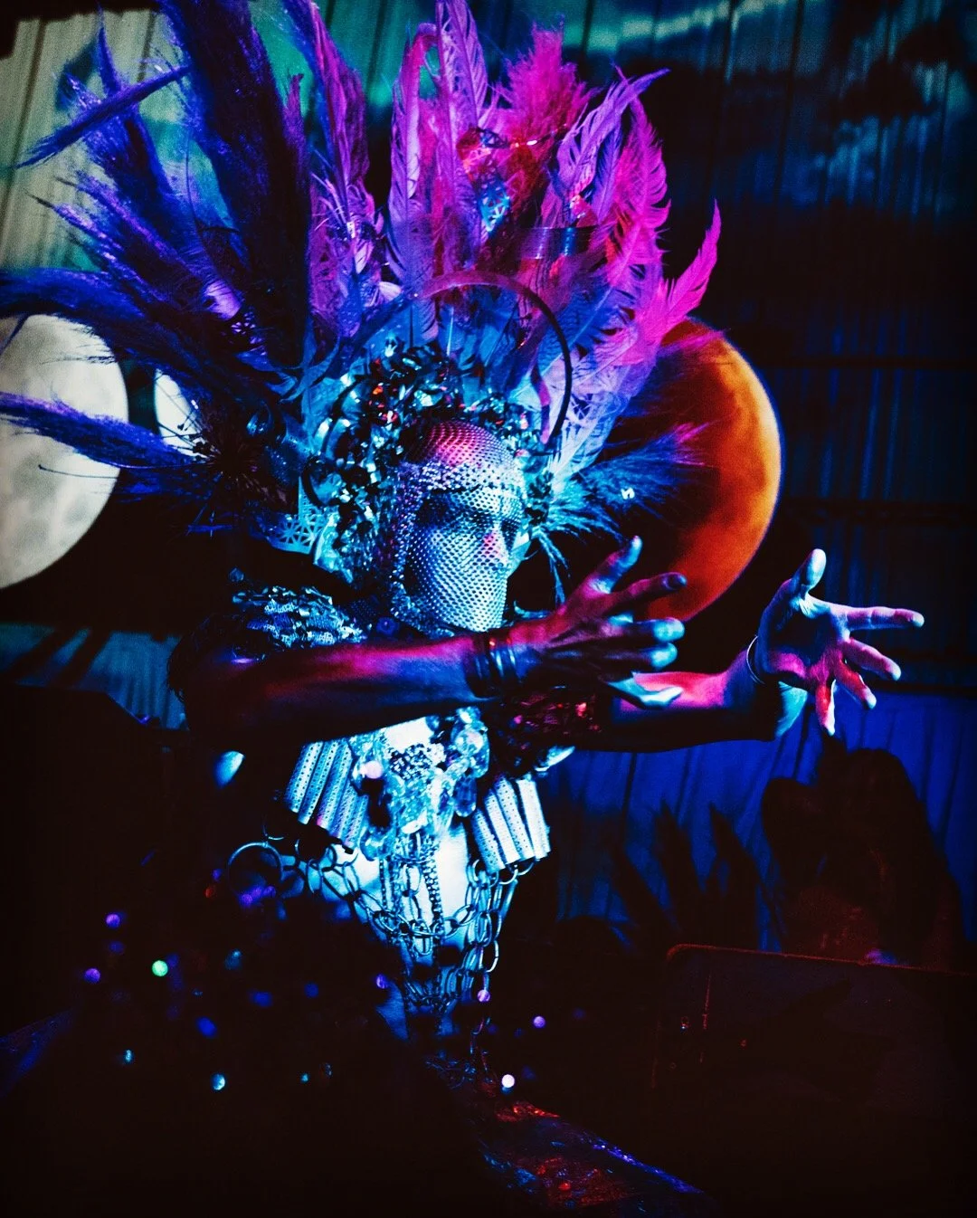 A performer wearing an elaborate costume with colorful feathers, rhinestones, and jewelry, dancing with arms extended in a vibrant, dark setting with multicolored lighting.