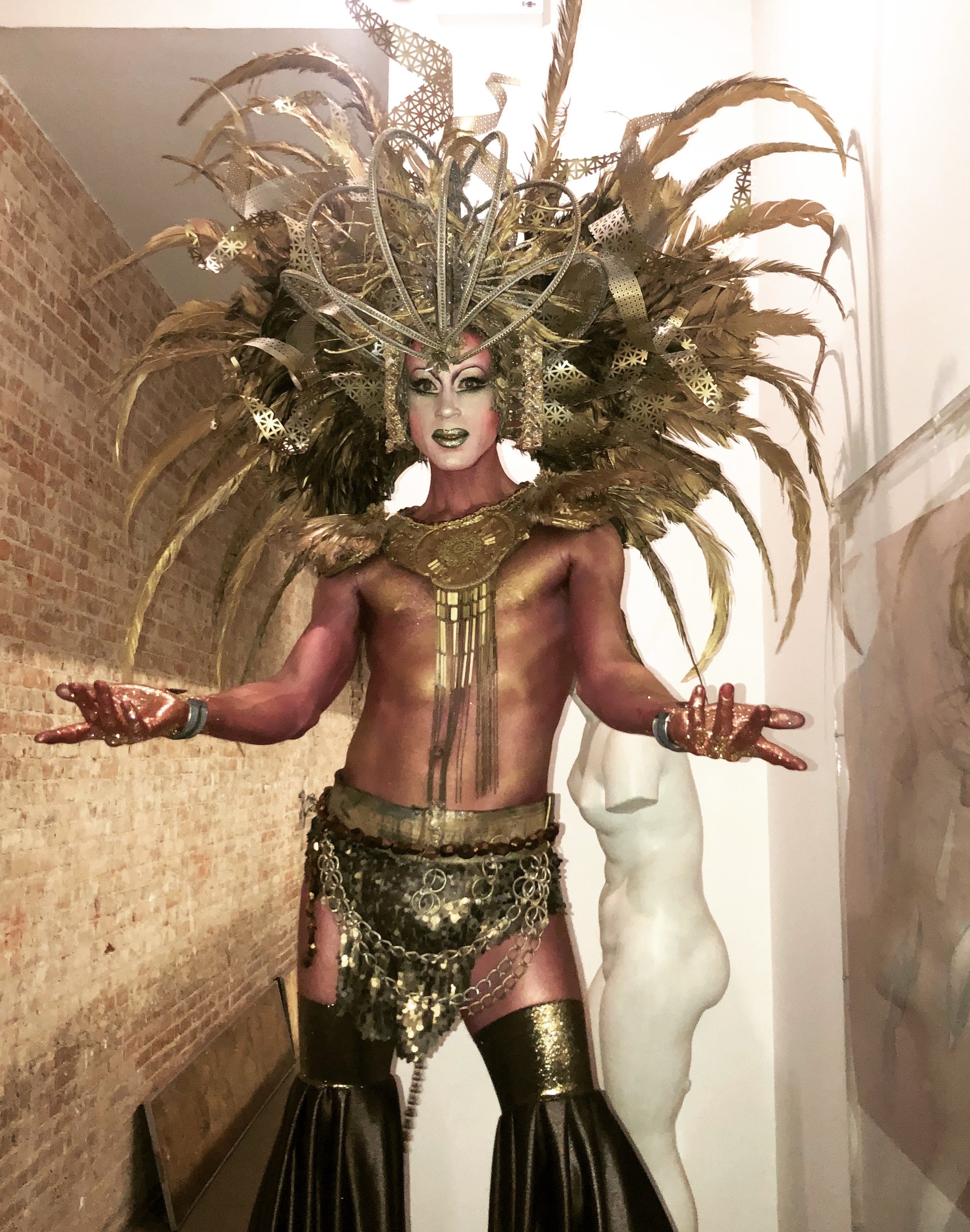 Performer in elaborate gold and black costume with large feathered headdress, gold makeup, and jewelry.
