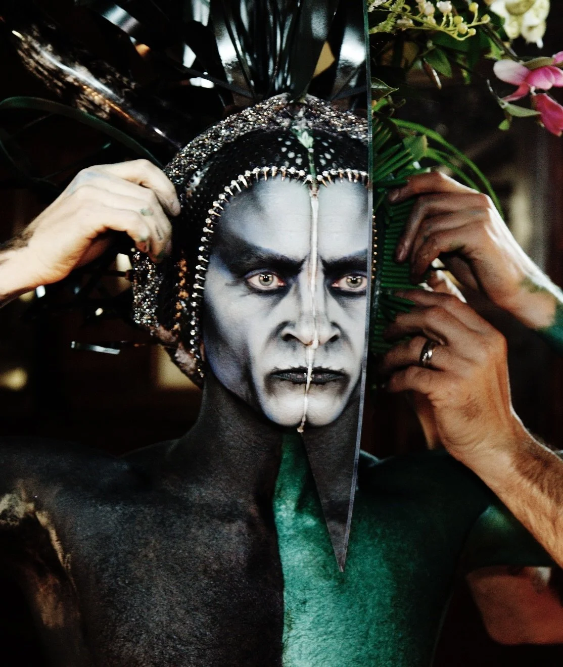 Person with elaborate face makeup, wearing an ornate headdress, holding a mirror, and dressed in green and black, with hands adjusting the headdress.