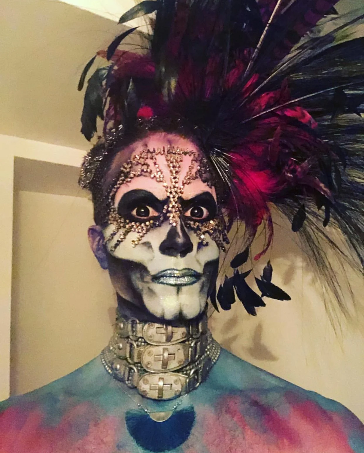 Person with elaborate Day of the Dead style face paint, wearing a headdress with black and pink feathers, and adorned with rhinestones and jewelry.
