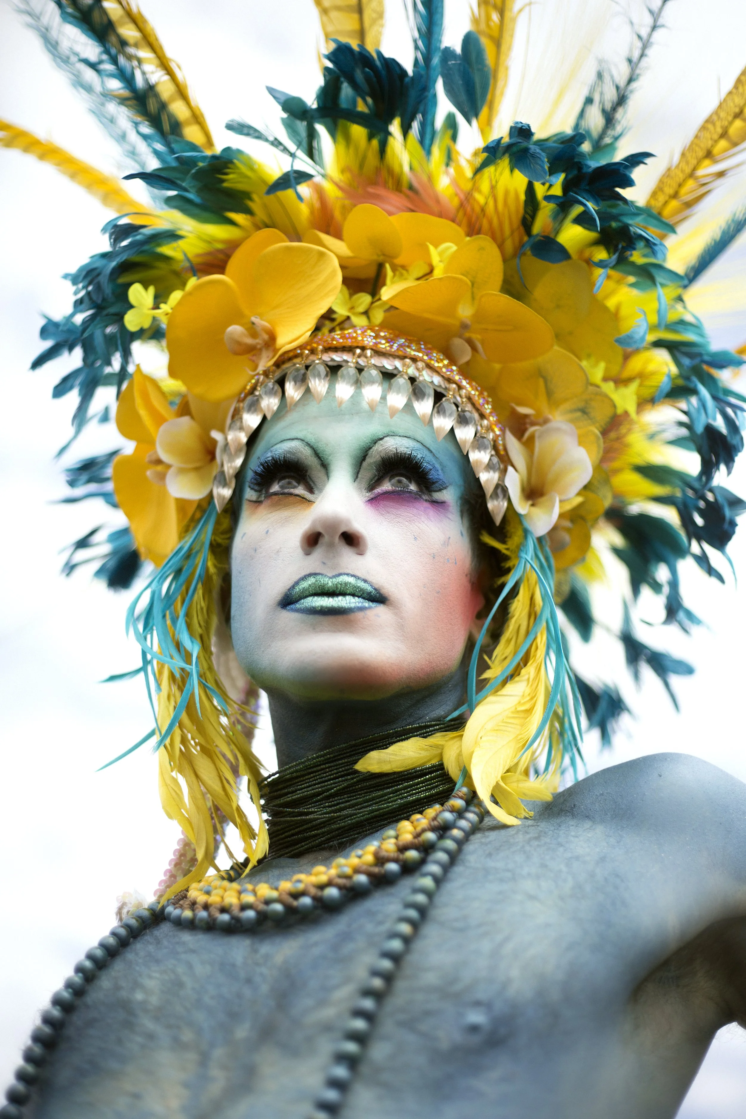 Person with elaborate colorful headdress made of yellow and blue feathers and flowers, vibrant makeup including green lipstick and multicolored eye shadow, looking upward.