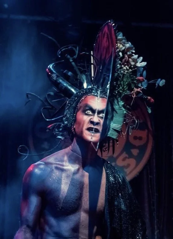 A performer dressed in elaborate, dark, fantasy-inspired costume with a headpiece adorned with large horns, feathers, flowers, and twisted metal. The individual has striking face makeup and an intense expression.
