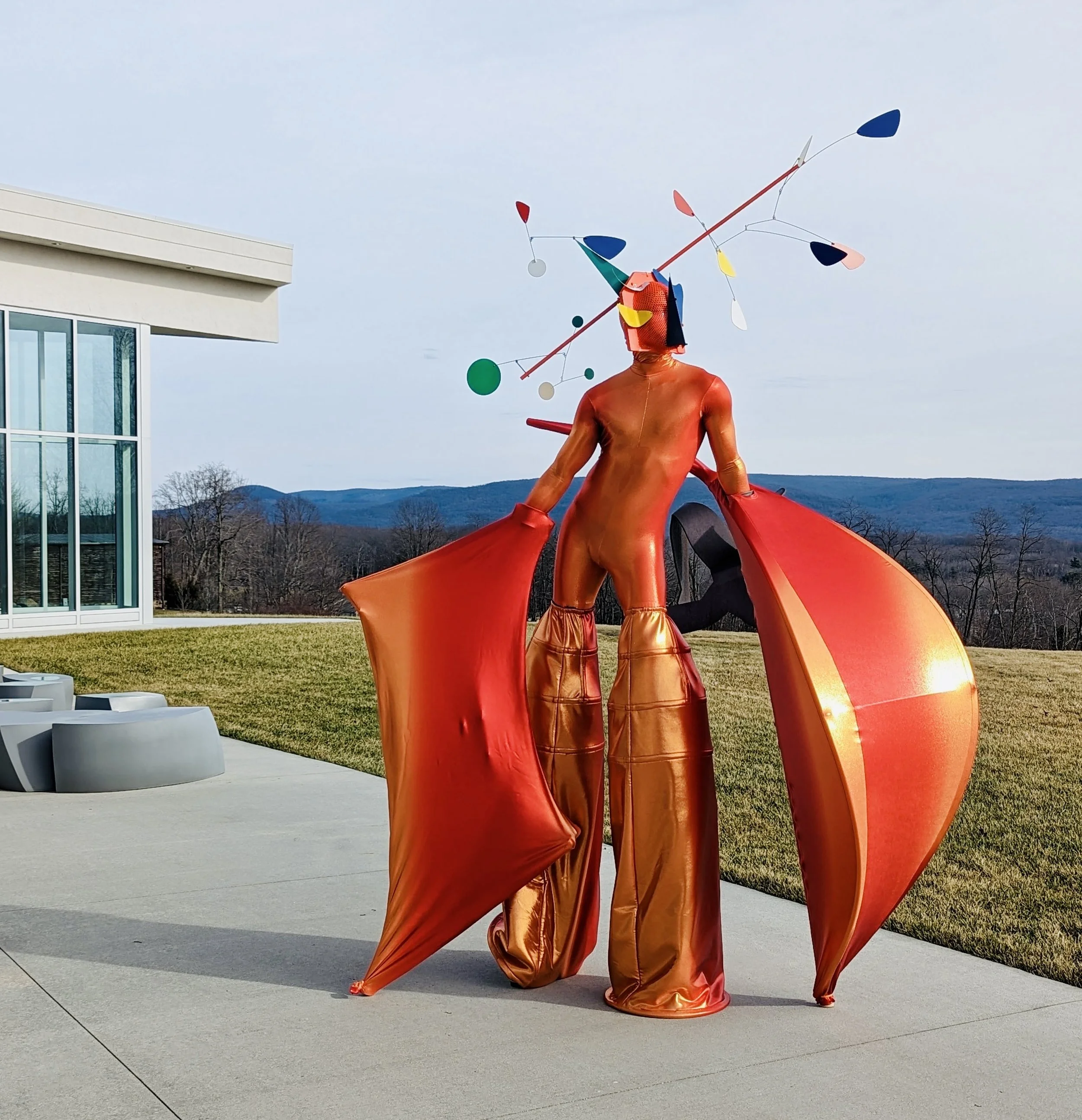 Colorful modern art sculpture of a figure with a metallic orange body, large red wings, and a head with a metal structure with hanging colorful shapes, situated outdoors near a building with glass windows and grassy landscape in the background.