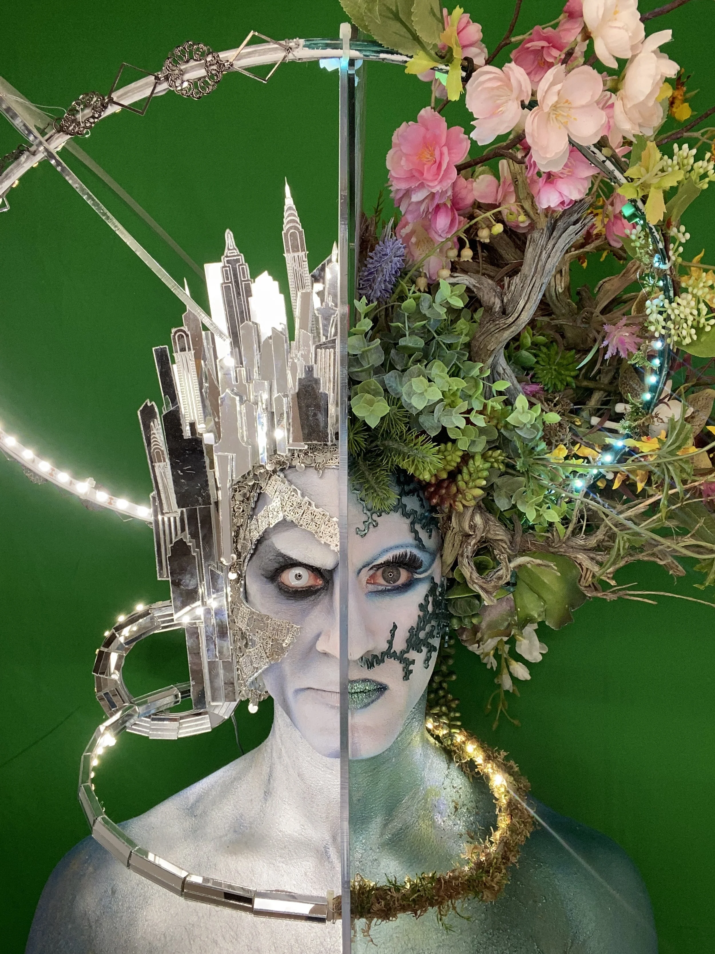 A split face sculpture of a woman, with the left side featuring a cityscape and silver jewelry, and the right side adorned with floral elements, greenery, and makeup, set against a green background.