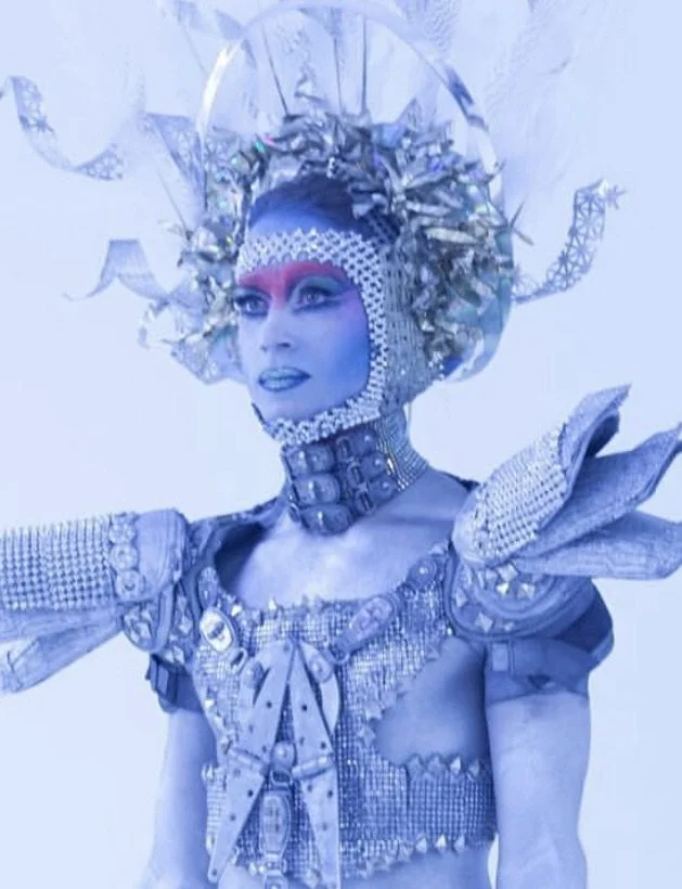 Person dressed in elaborate, futuristic costume with metallic details, silver and blue makeup, and a large headpiece with metallic and reflective elements.