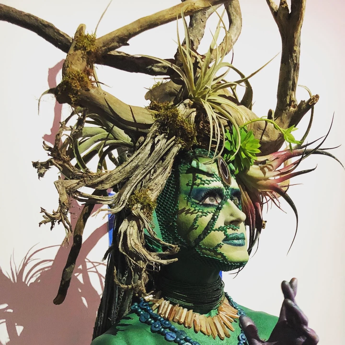 A person with elaborate green and blue face paint, wearing tribal-style jewelry, and a headdress made of intertwined driftwood, plants, and moss.