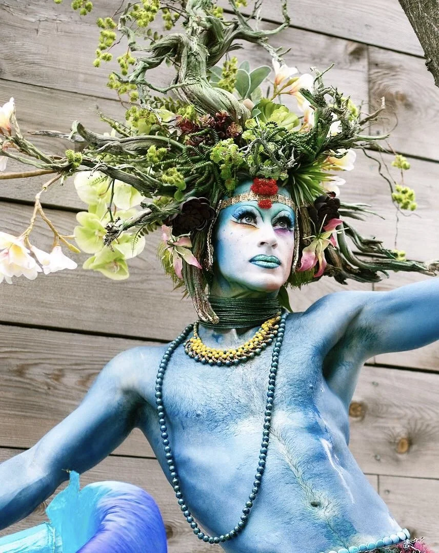 A person with blue body paint and elaborate headdress made of tree branches, flowers, and greenery, styled in a fantasy or artistic costume, against a wooden wall background.