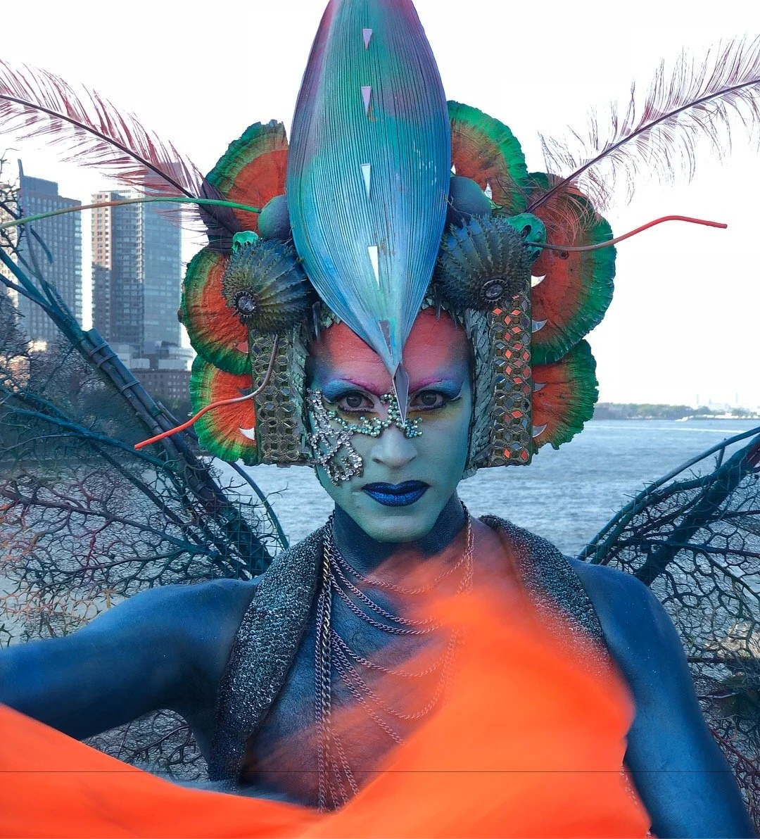 Person wearing elaborate, colorful costume with feathers and intricate face makeup, posing outdoors near water with cityscape in the background.