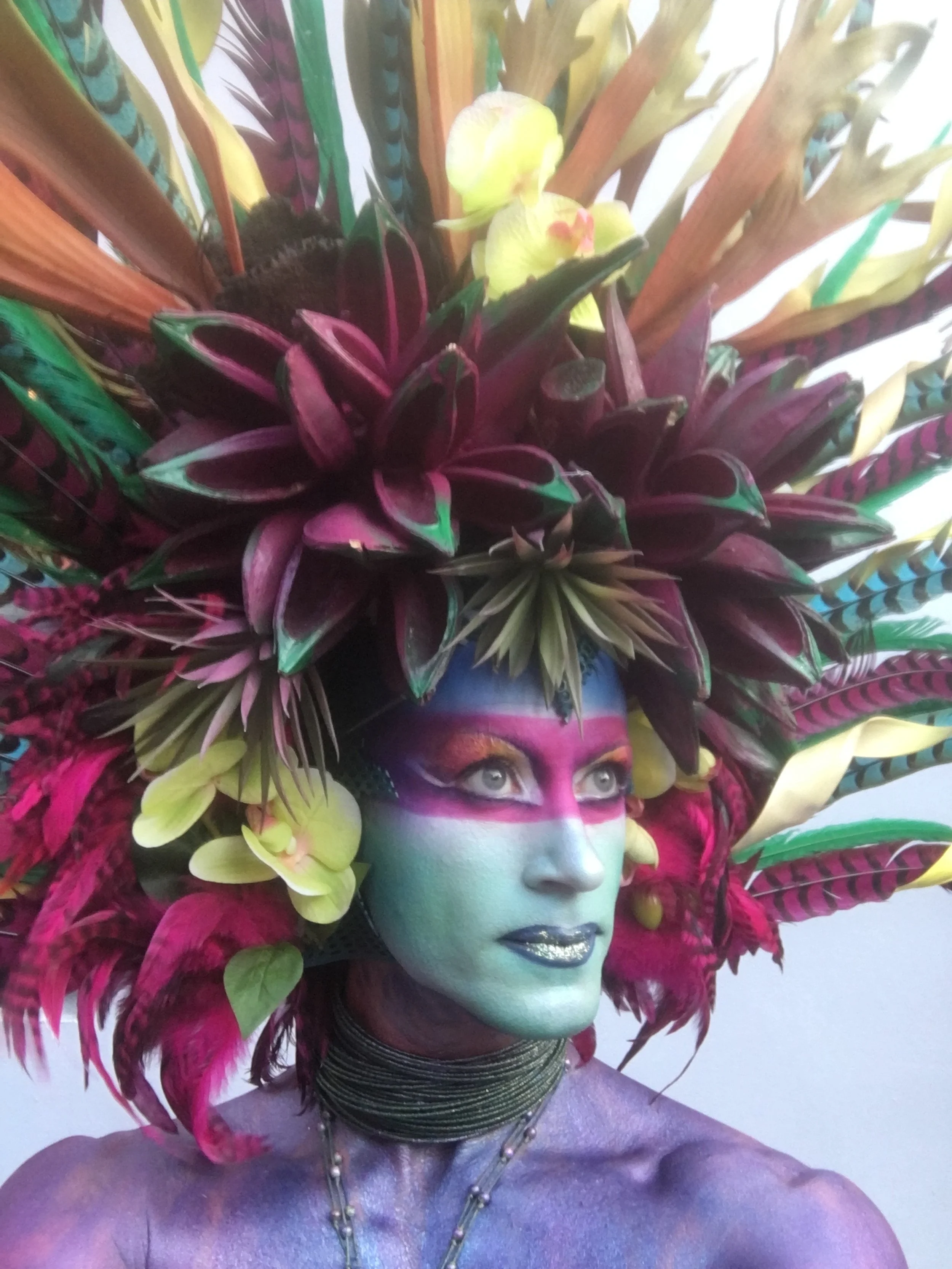Person with vibrant, colorful makeup and an elaborate headdress made of various tropical and exotic flowers and leaves.