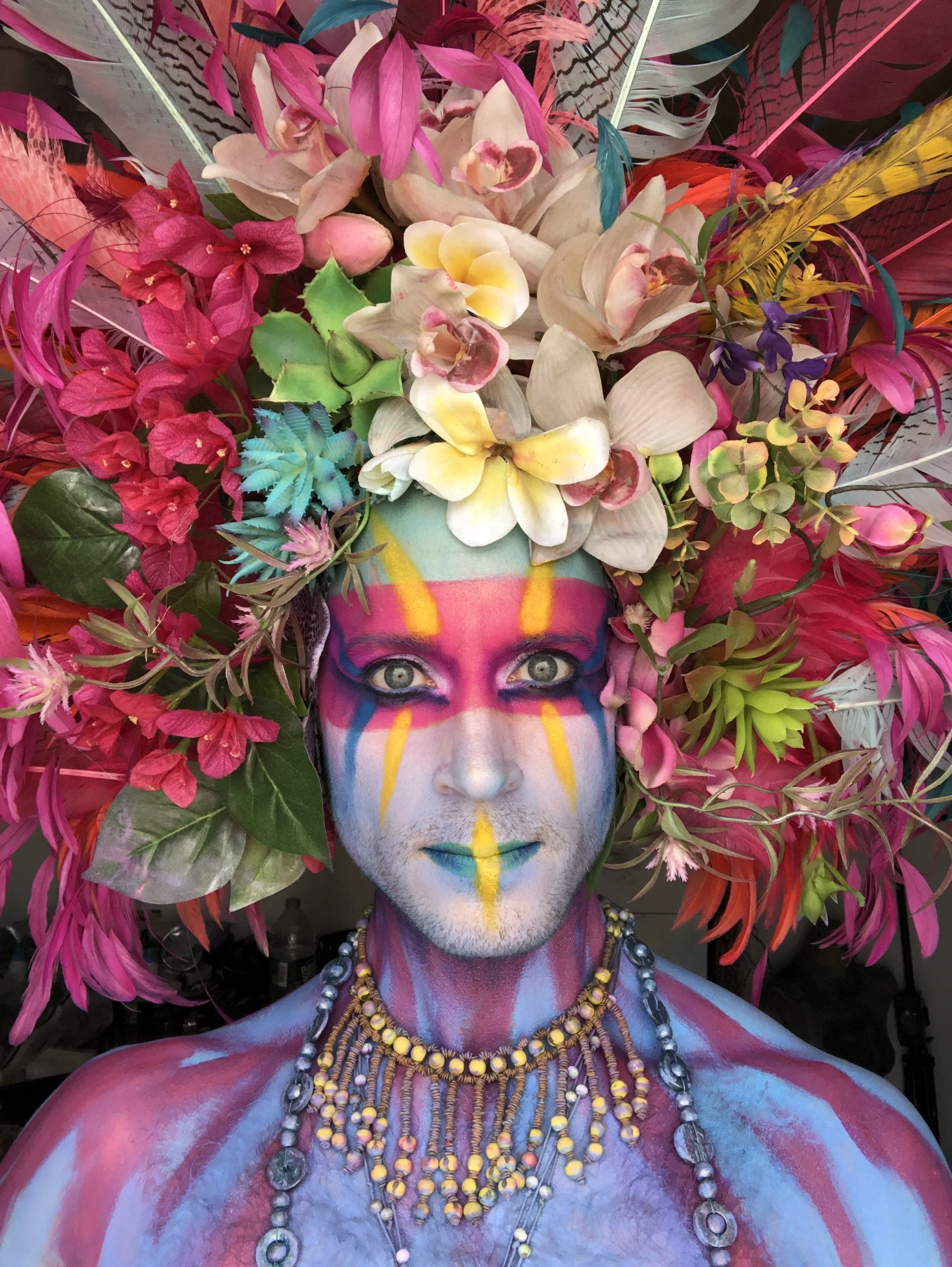Person with elaborate colorful body paint, wearing a large headdress made of pink, white, yellow, and purple flowers, with a matching floral necklace, makeup with bright colors and streaks, and staring directly at the camera.