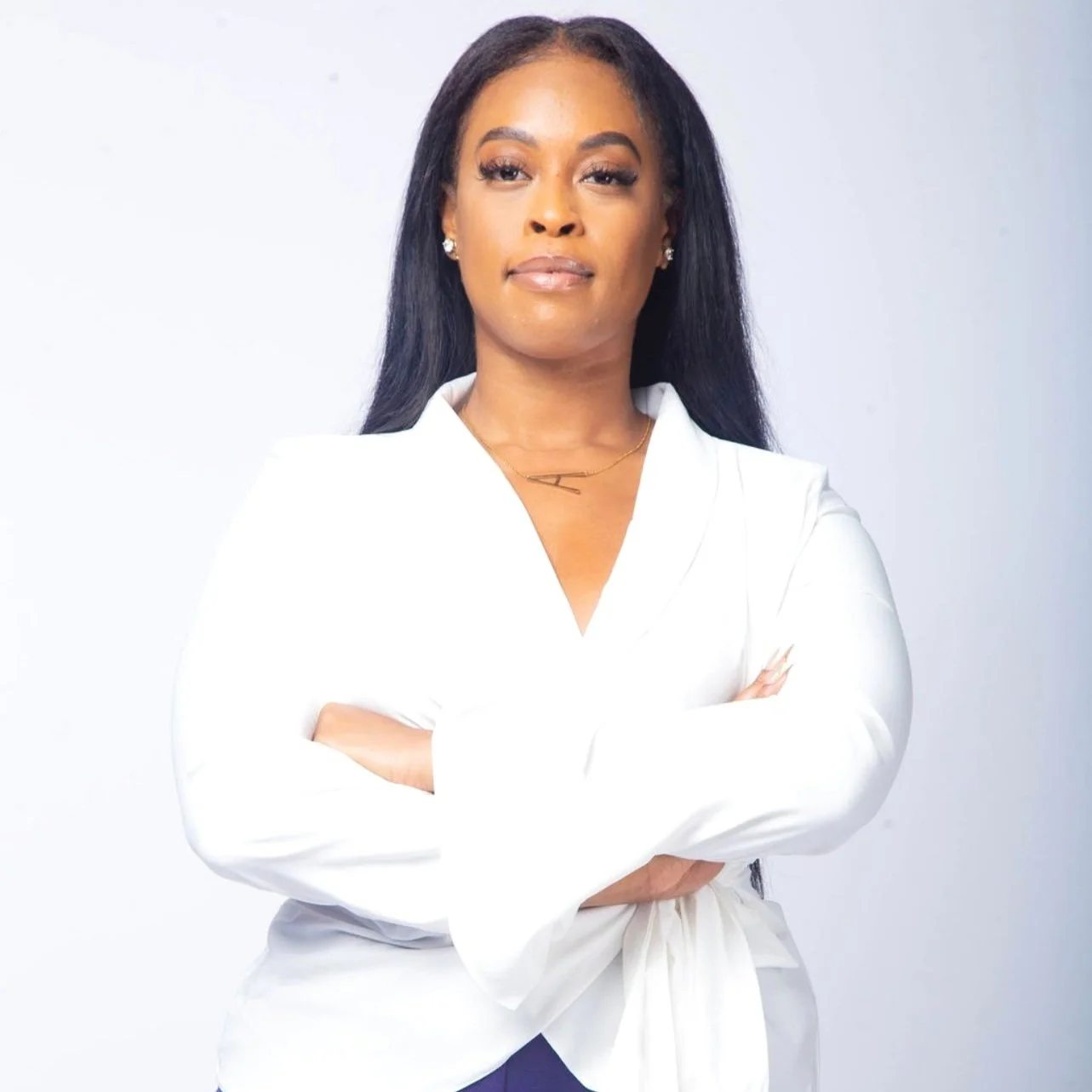 A confident woman with medium skin tone and long black hair, wearing a white blazer with arms crossed, stands against a plain light background.