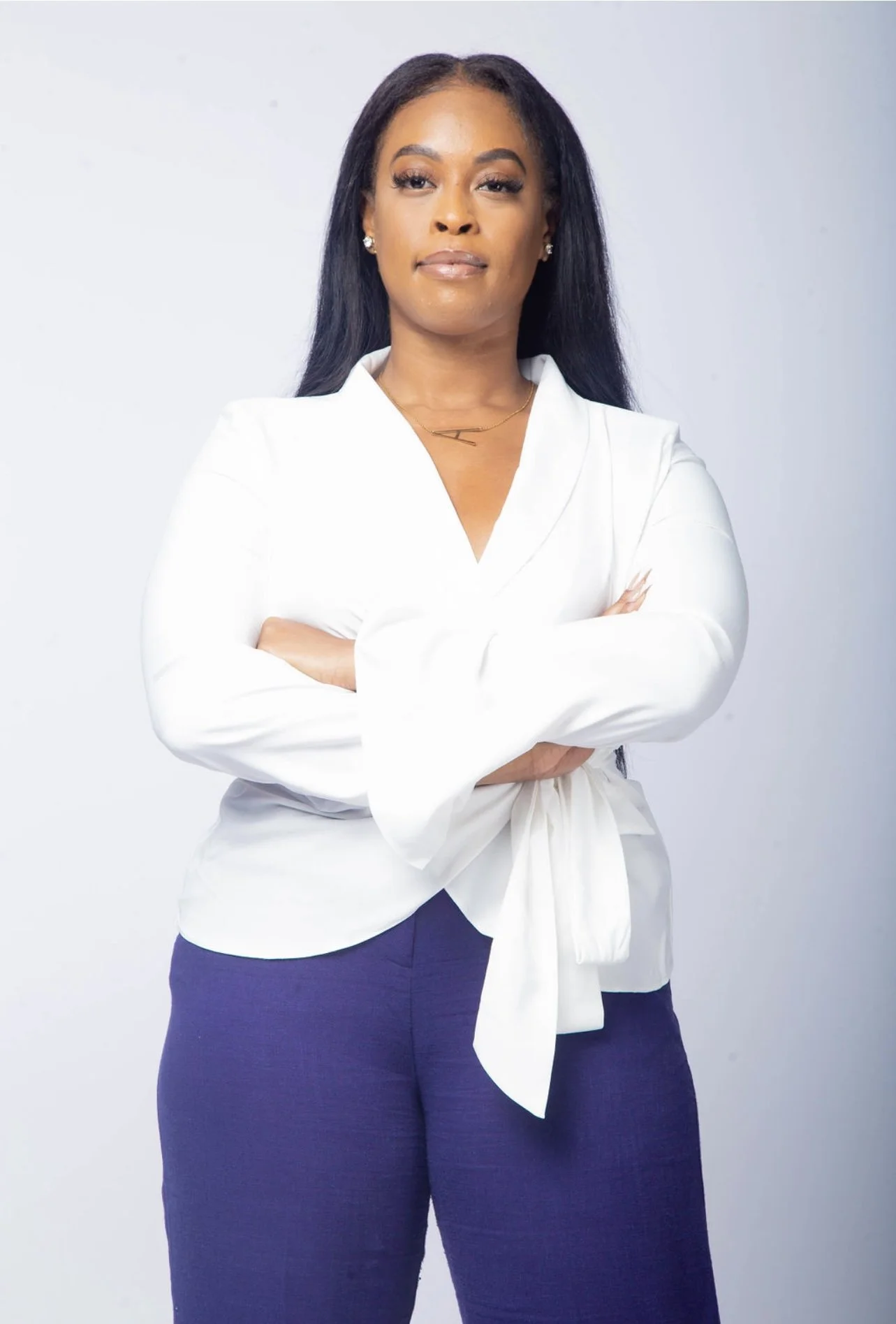 A woman with medium-dark skin, long black hair, and wearing a white blazer with a tied waist, dark blue pants, and jewelry, stands with arms crossed against a light background.
