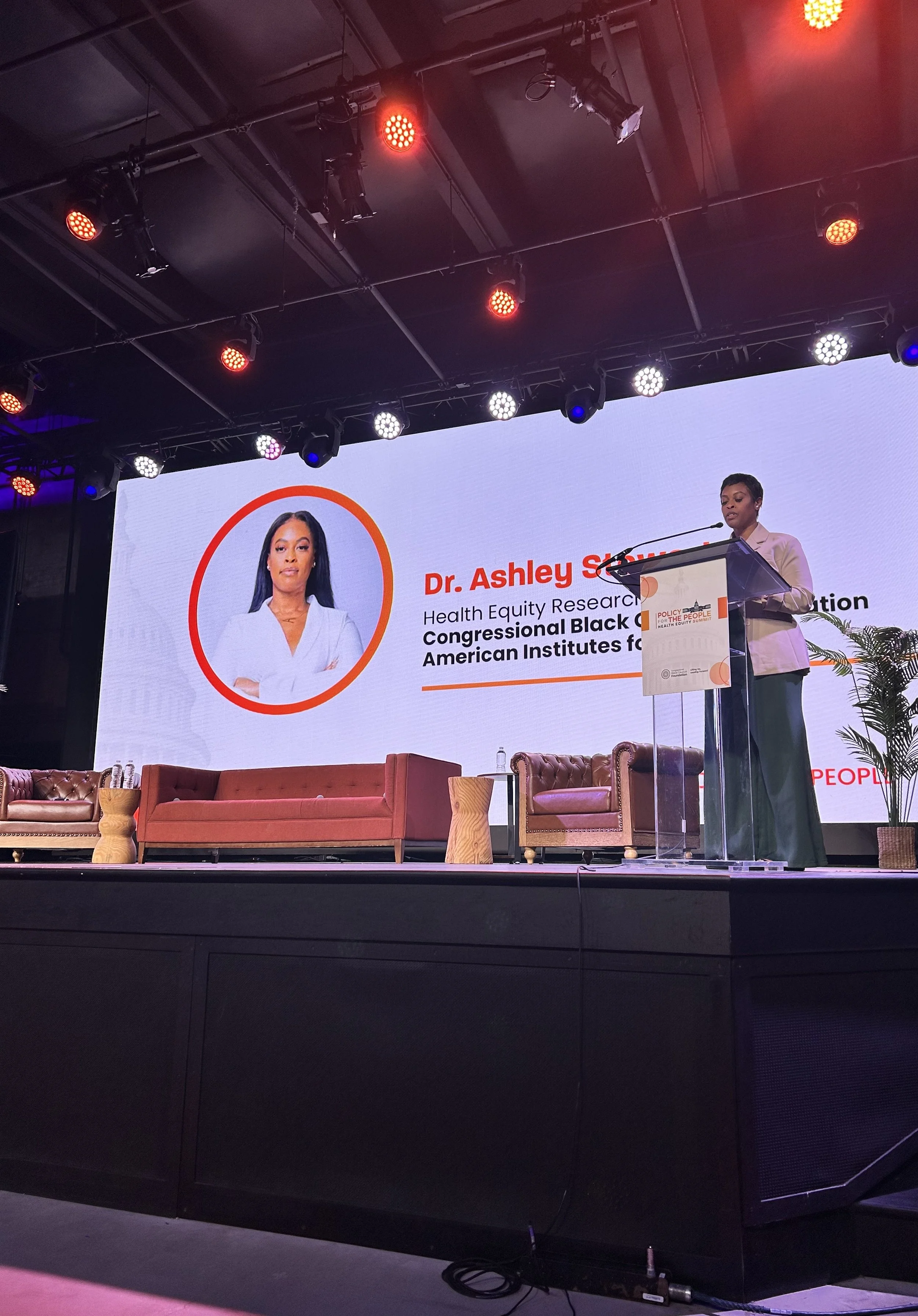Dr. Ashley M. Stewart is speaking at a podium. The slide includes her name and titles related to health equity research and congressional black organizations. There are couches and chairs on the stage, with lighting and equipment overhead.
