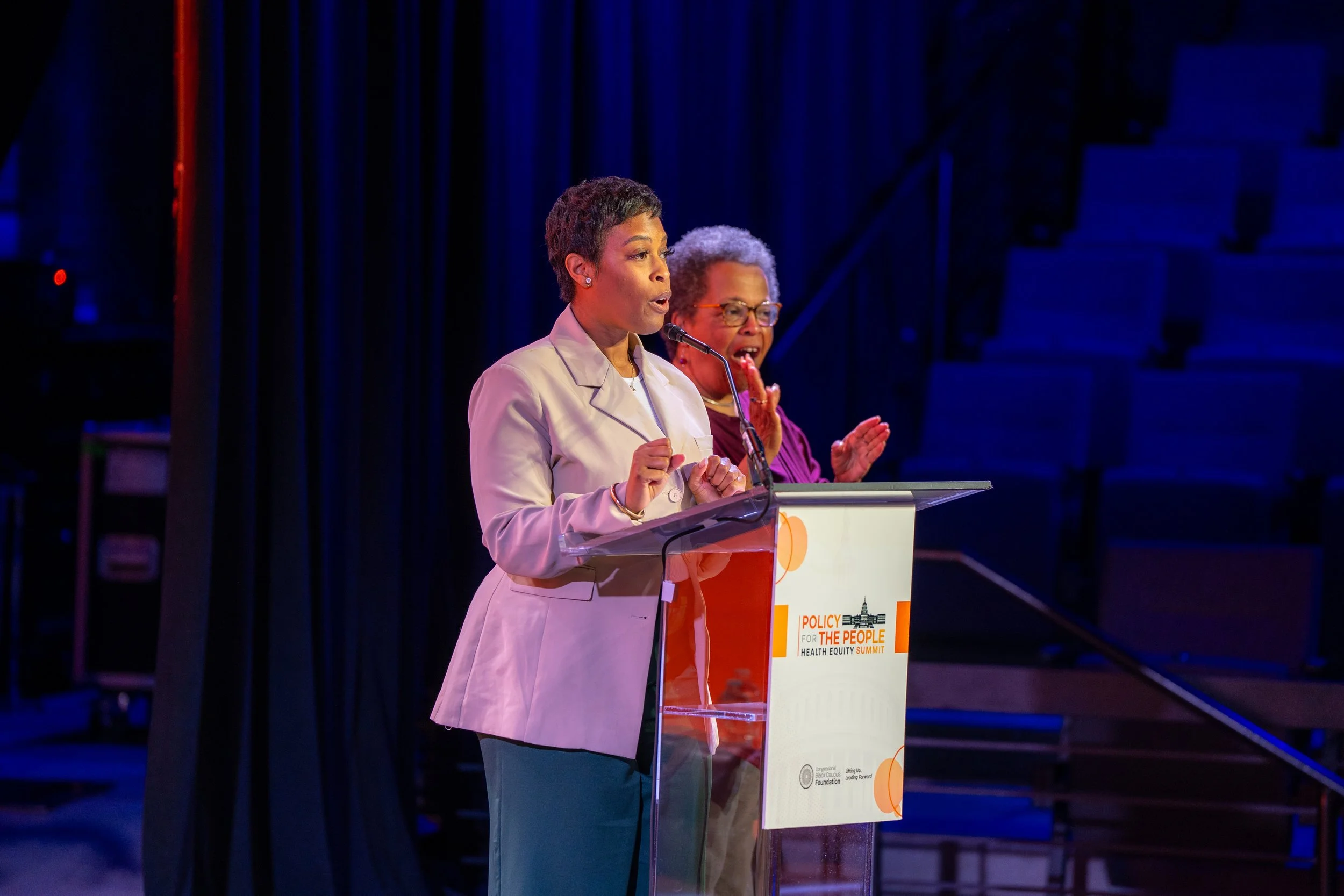 Two women at a podium speaking at a conference. One woman is wearing a light beige blazer and the other is in a purple outfit. The podium has a sign with text about health equity summit.