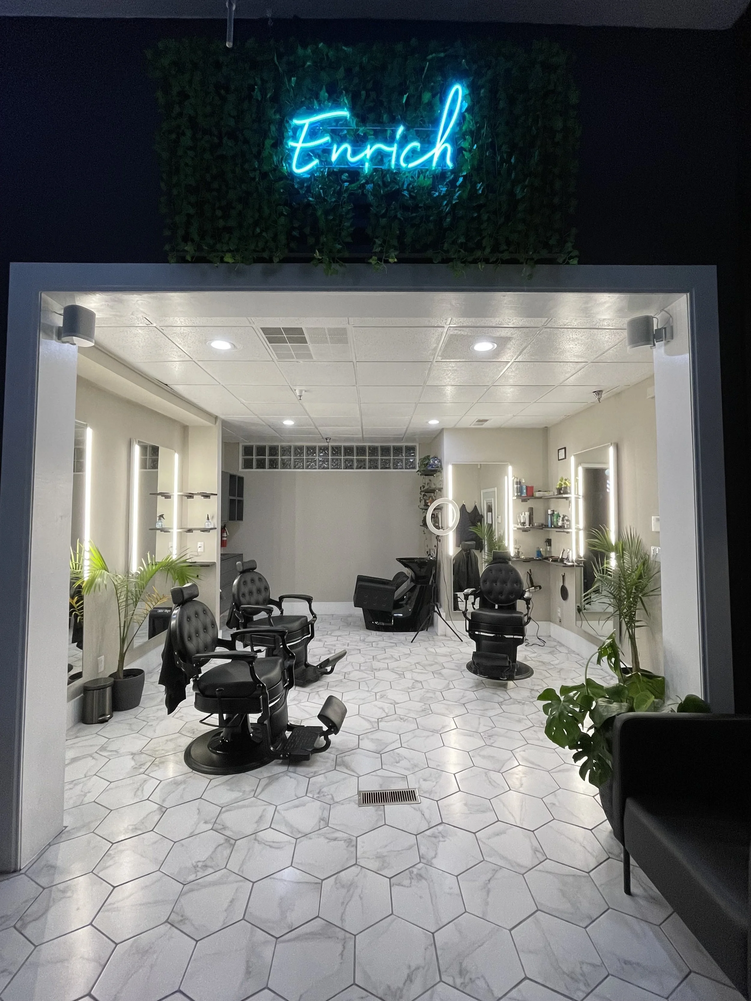 Interior view of a modern barbershop with black reclining barber chairs, illuminated mirrors, potted plants, shelves with grooming products, and a neon sign reading 'Enrich' above the entrance.
