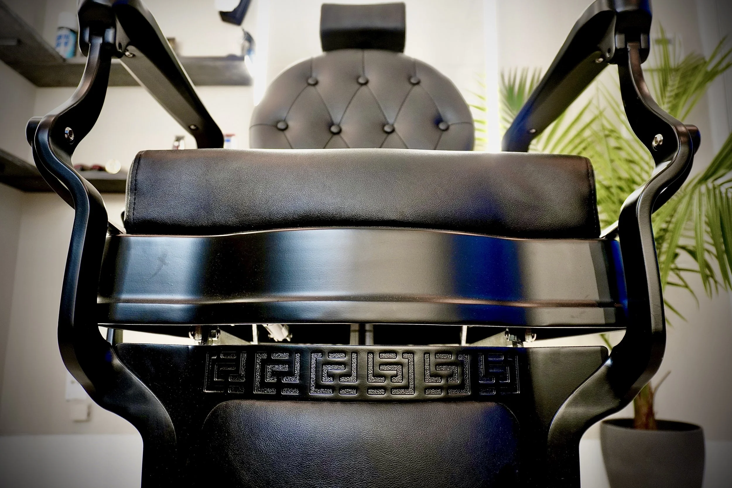 Front view of a black barber chair with a padded headrest, leather seat, and decorative cut-out pattern on the back, with a plant and shelves in the background.