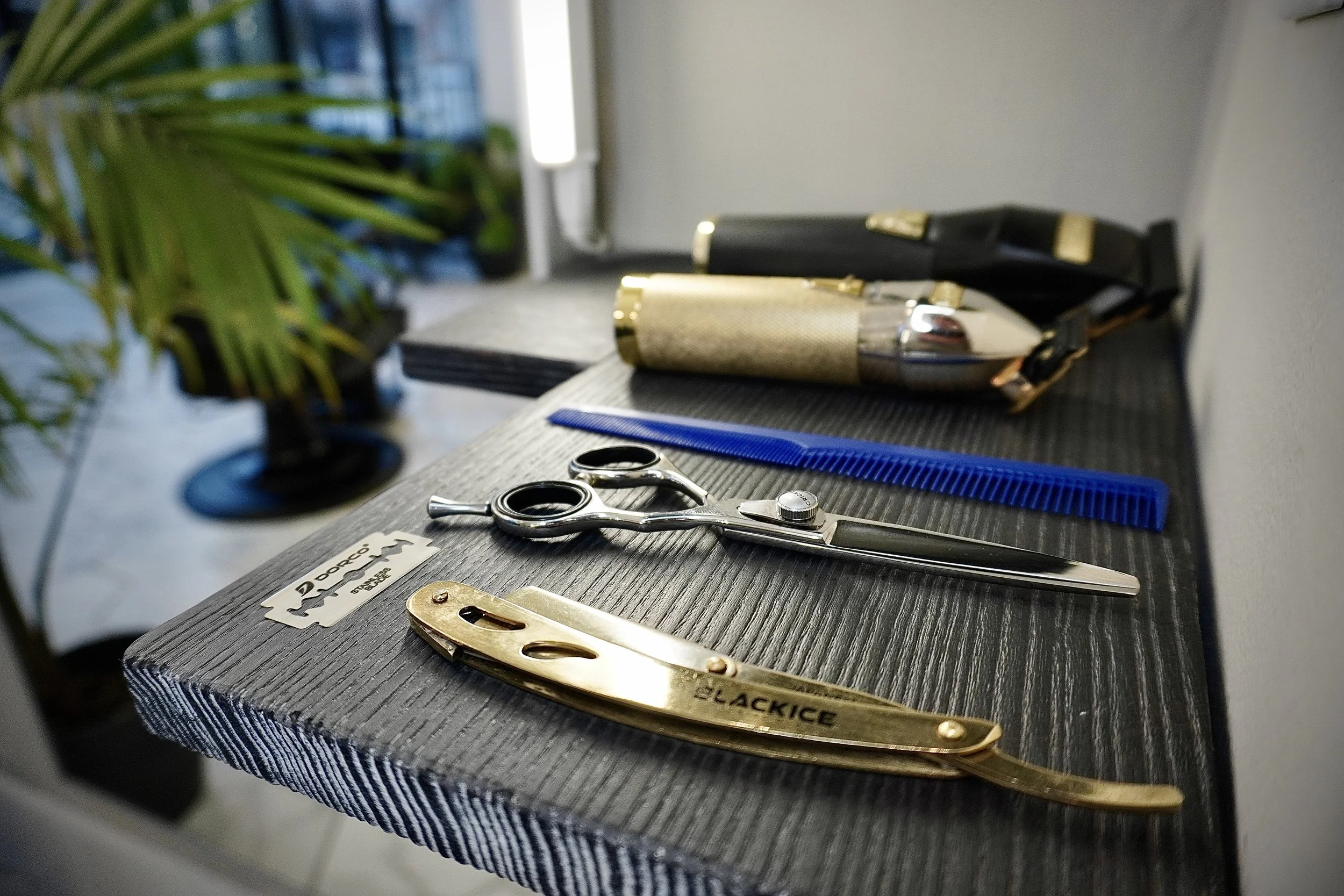 Hairdressing tools including scissors, a blue comb, a gold hair clipper, and a spray bottle on a black wooded surface with some spray bottles and a plant in the background.