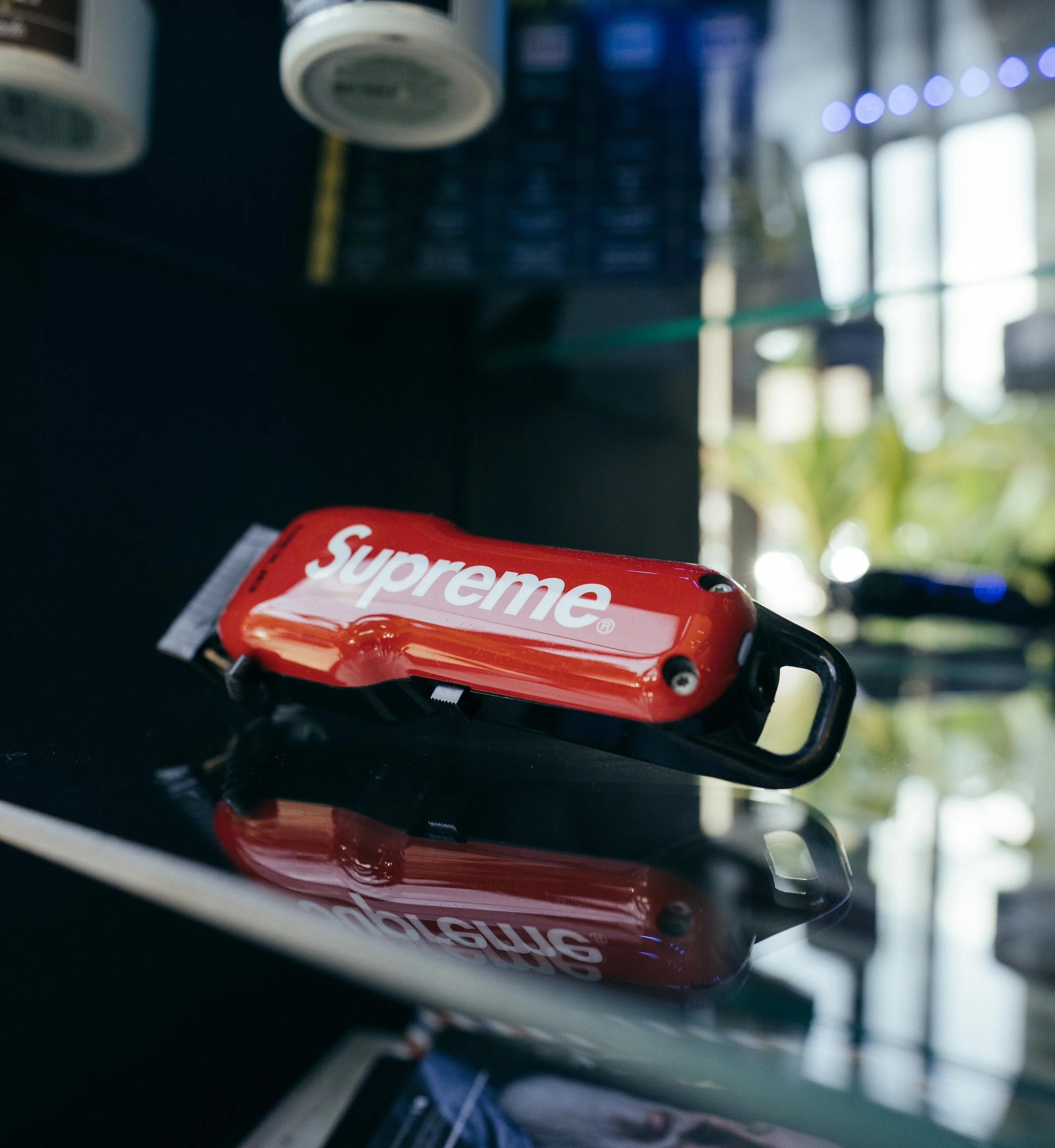 A red Supreme branded bicycle lock resting on a reflective black surface with a blurred background of indoor and outdoor elements.