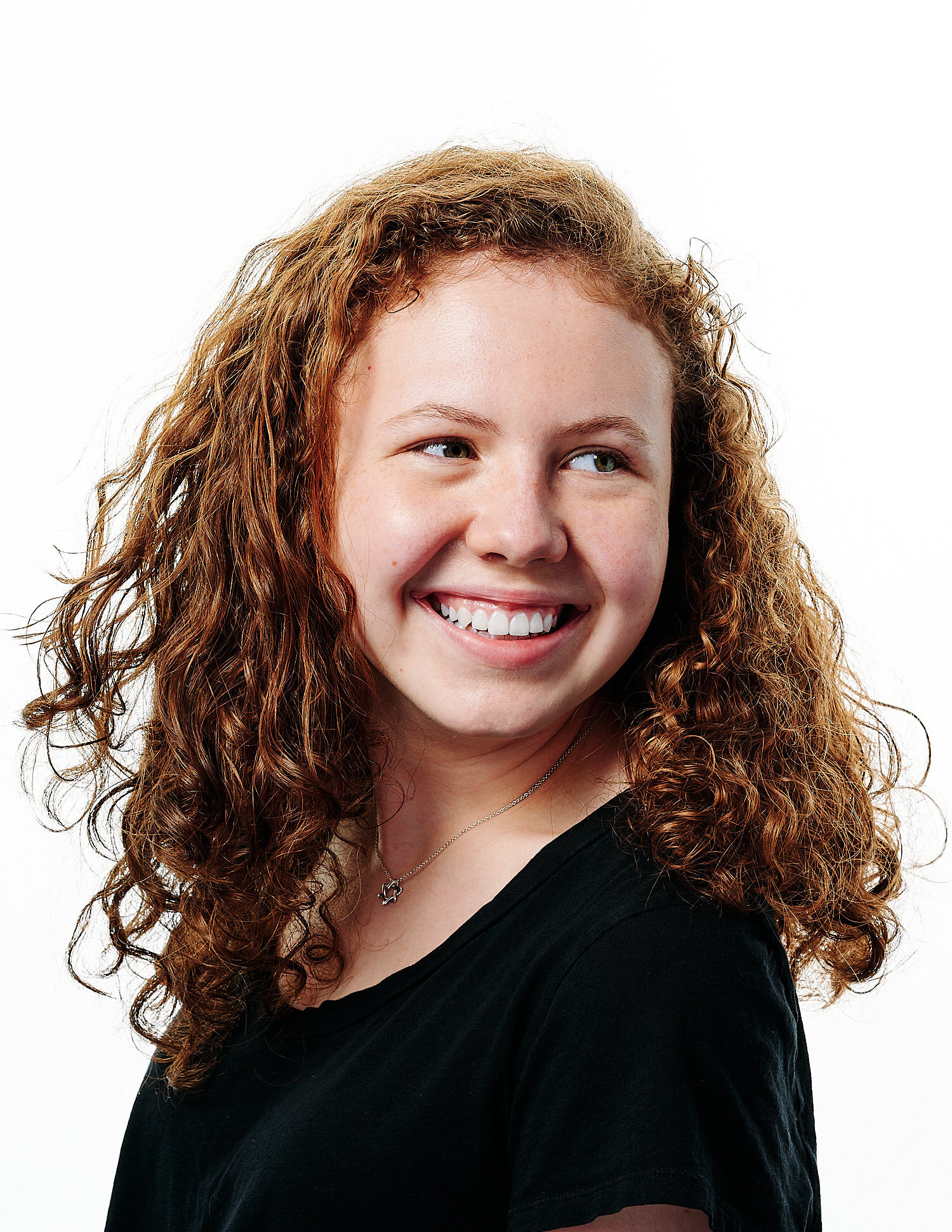  teenage actor posing for a headshot picture in a professional photo studio with white background. She is caucasian, has curly red hair, a round freckled face with full lips. She wears black t-shirt. 