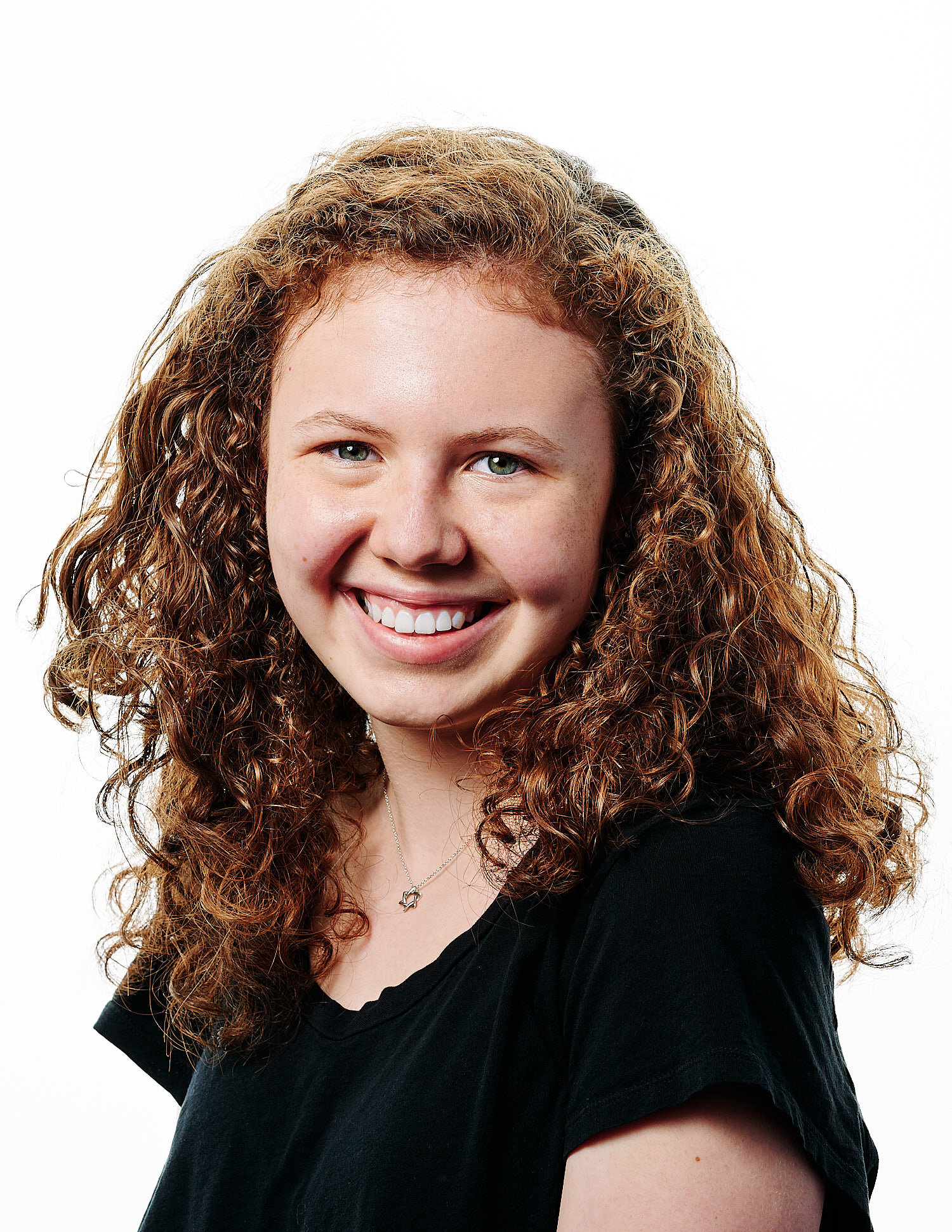  teenage actor posing for a headshot picture in a professional photo studio with white background. She is caucasian, has curly red hair, a round freckled face with full lips. She wears black t-shirt. 