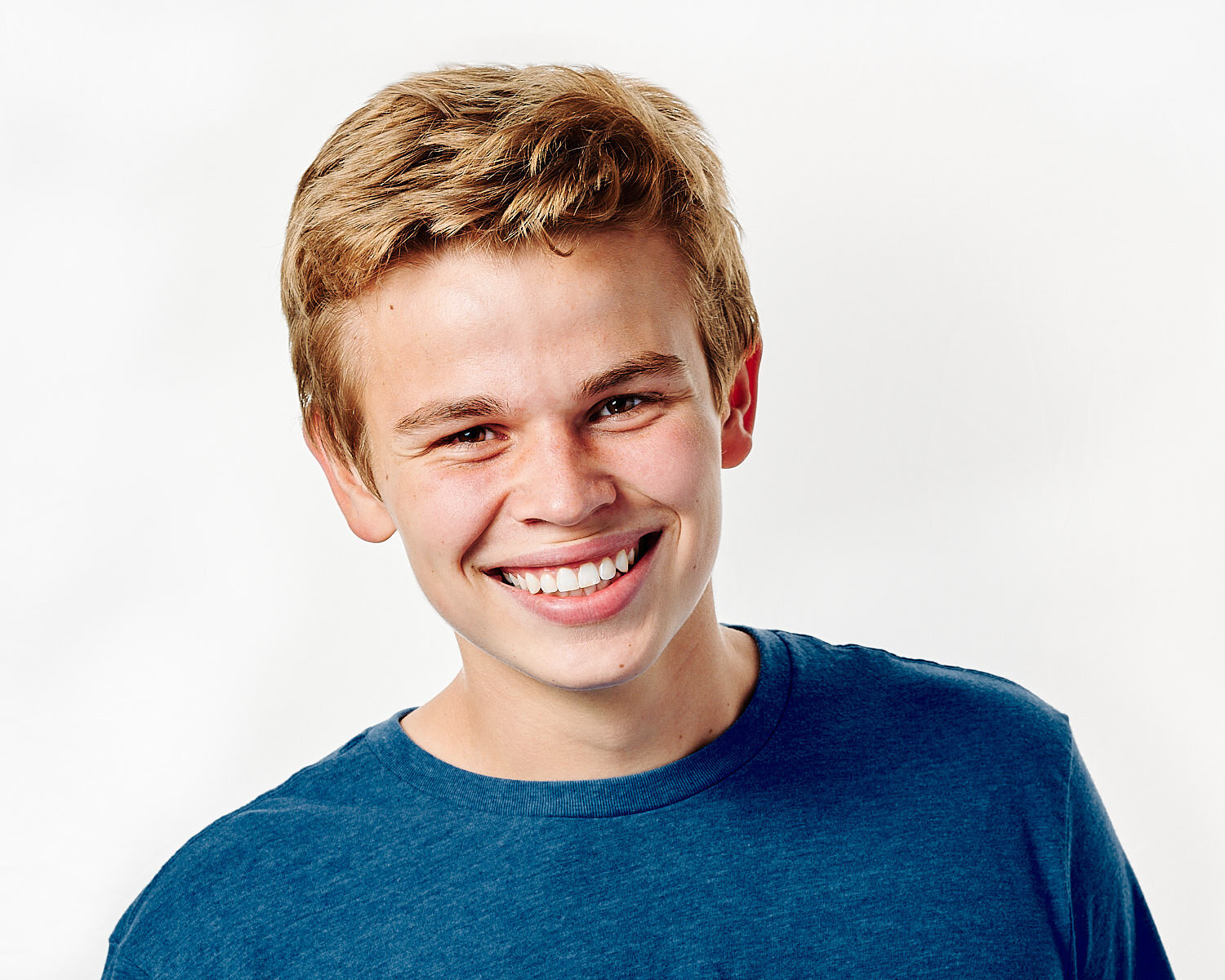  Max Peluso is posing for actor headshots against white background in a professional photography studio. He looks handsome and happy, he has blond hair and wears a blue t-shirt. 