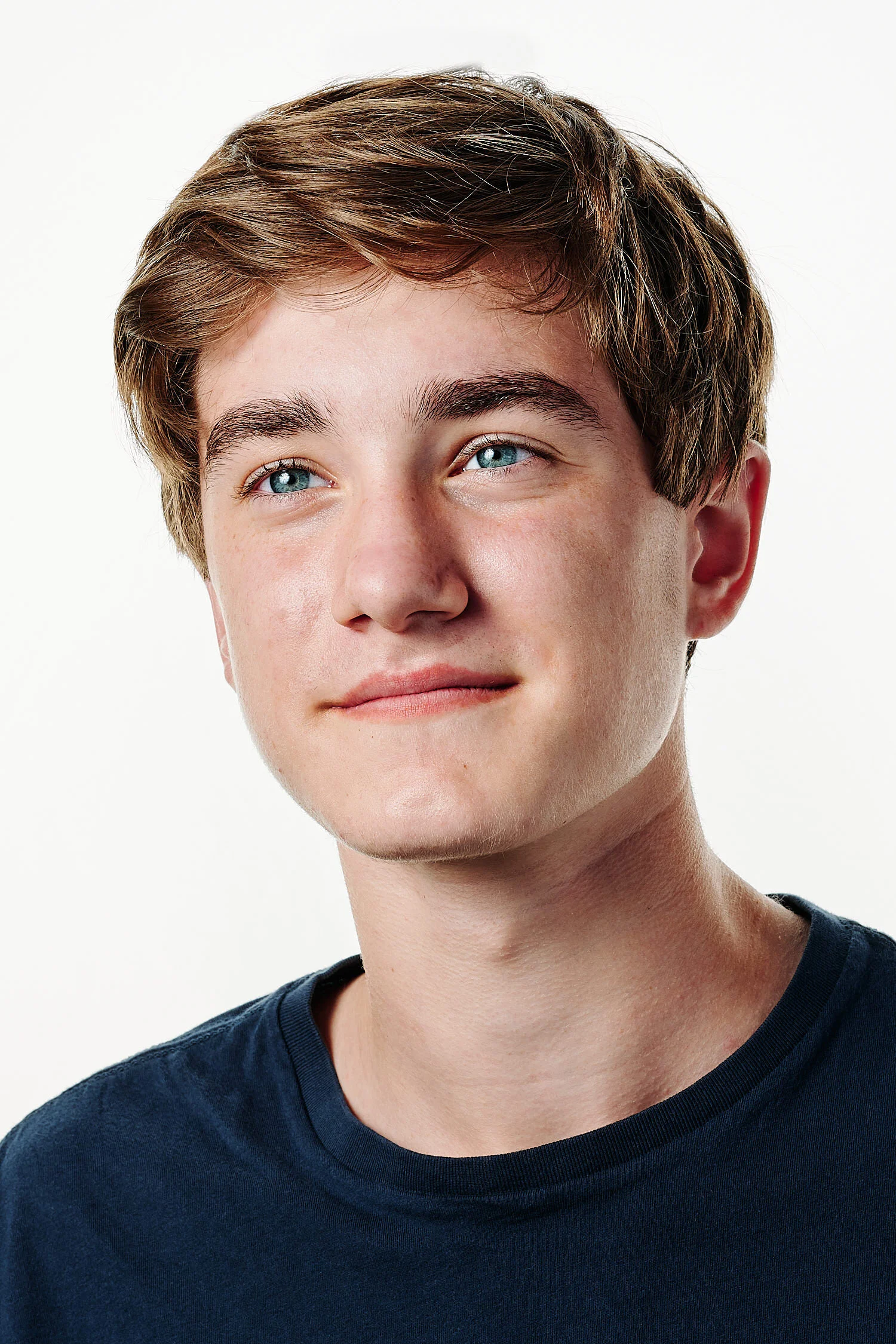  Logan Carlson is posing for actor headshots against white background in a professional photography studio. He looks handsome and happy, he has fair hair and wears a navy t-shirt. 