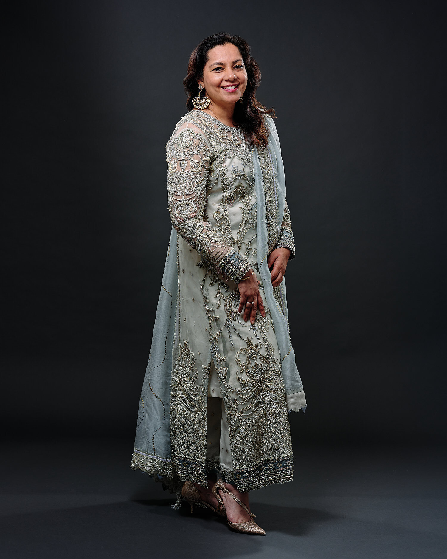  Bushra and Saad are posing for their wedding portraits in a professional photography studio. Bushra is dressed in a stunning pale dress ornamented with pearls, floral motives. Saad wears a blue suit. 